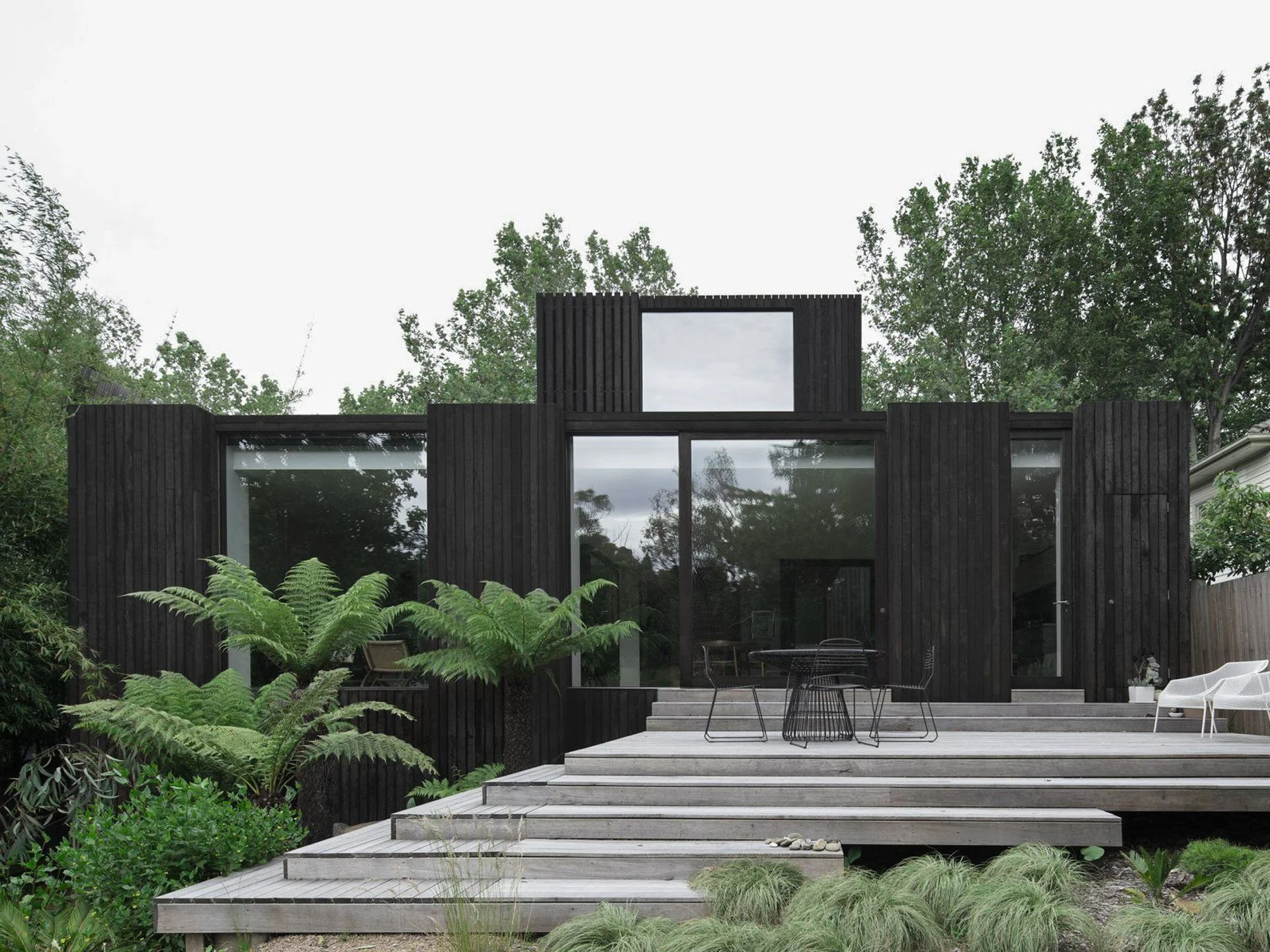 Modern black wooden house with large glass windows, silver stairs, and outdoor seating, surrounded by green trees and plants.