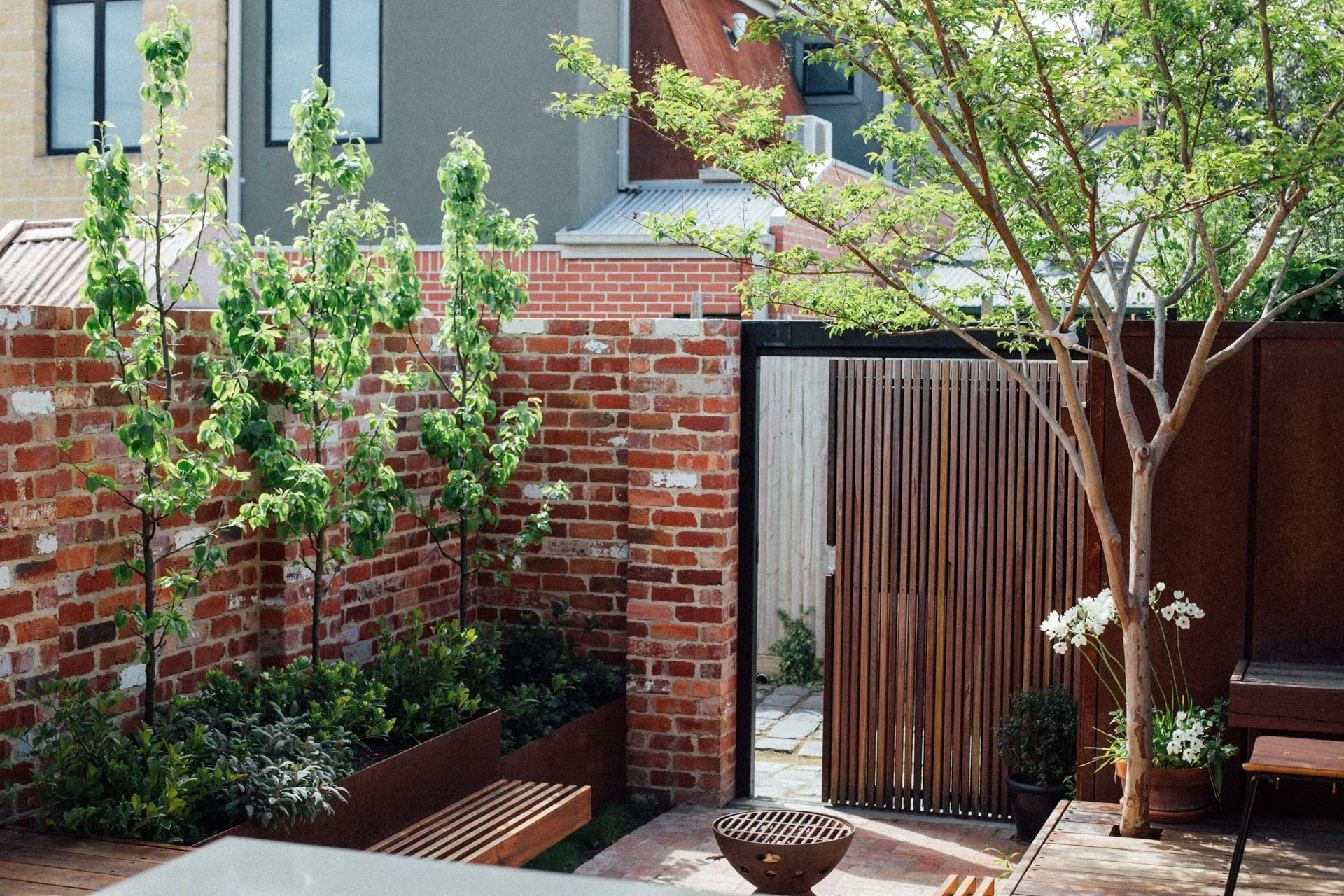 An outdoor garden area featuring a brick wall with greenery, a wooden gate, and trees with green leaves. There are potted plants with white flowers, a wooden bench, and a small firepit.