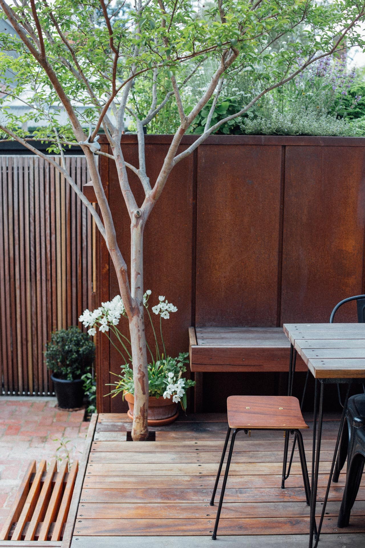 A cozy outdoor patio features a small wooden deck area with a tree, potted plants, a wooden bench, a table, and a stool, enclosed by a wooden fence and privacy panels, with greenery visible.