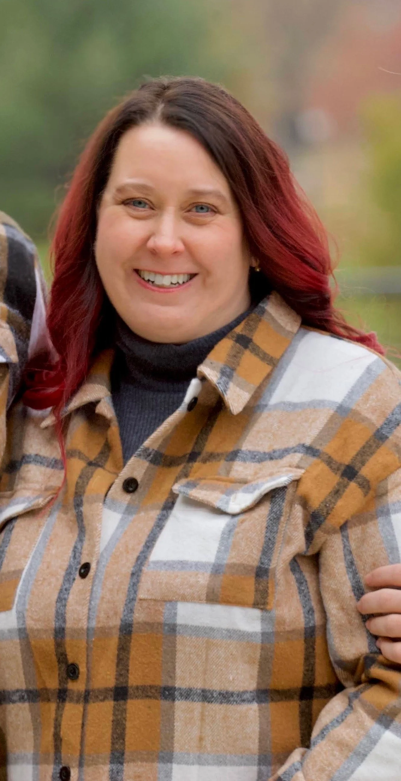 A woman with shoulder-length dark brown and red hair, smiling, wearing a black turtleneck and a tan, white, and black plaid flannel shirt, outdoors with blurred green and autumn-colored background.