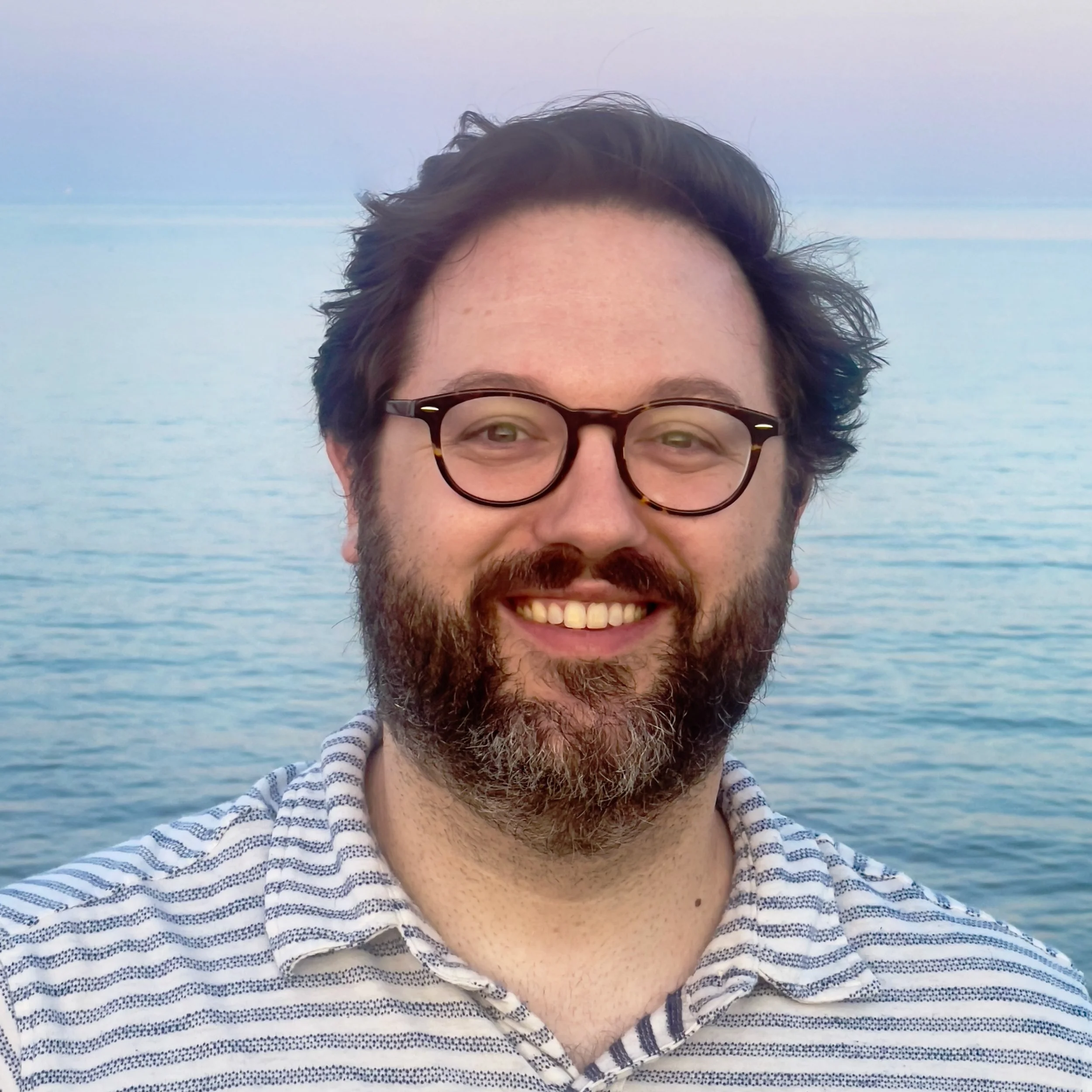 A smiling man with glasses and a beard, wearing a striped shirt, standing by a calm body of water during dusk or dawn.