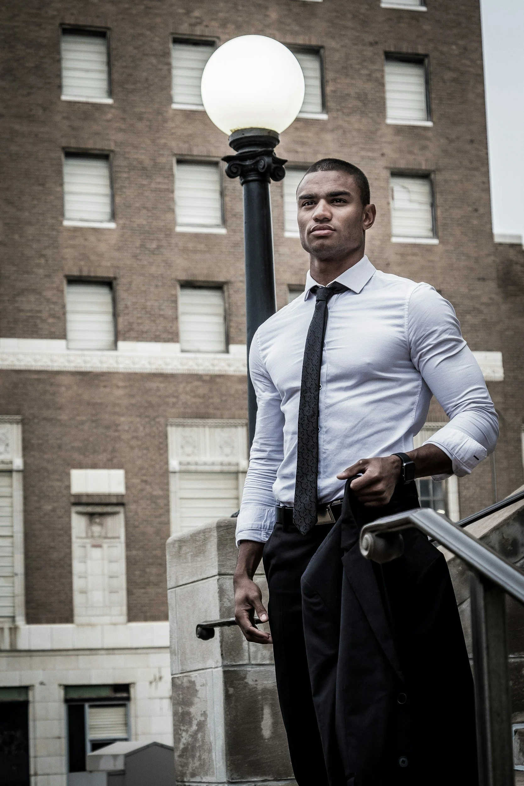A young man in business attire, white shirt, black tie, holding a black jacket, standing outdoors next to a lamppost in front of a brown brick building.
