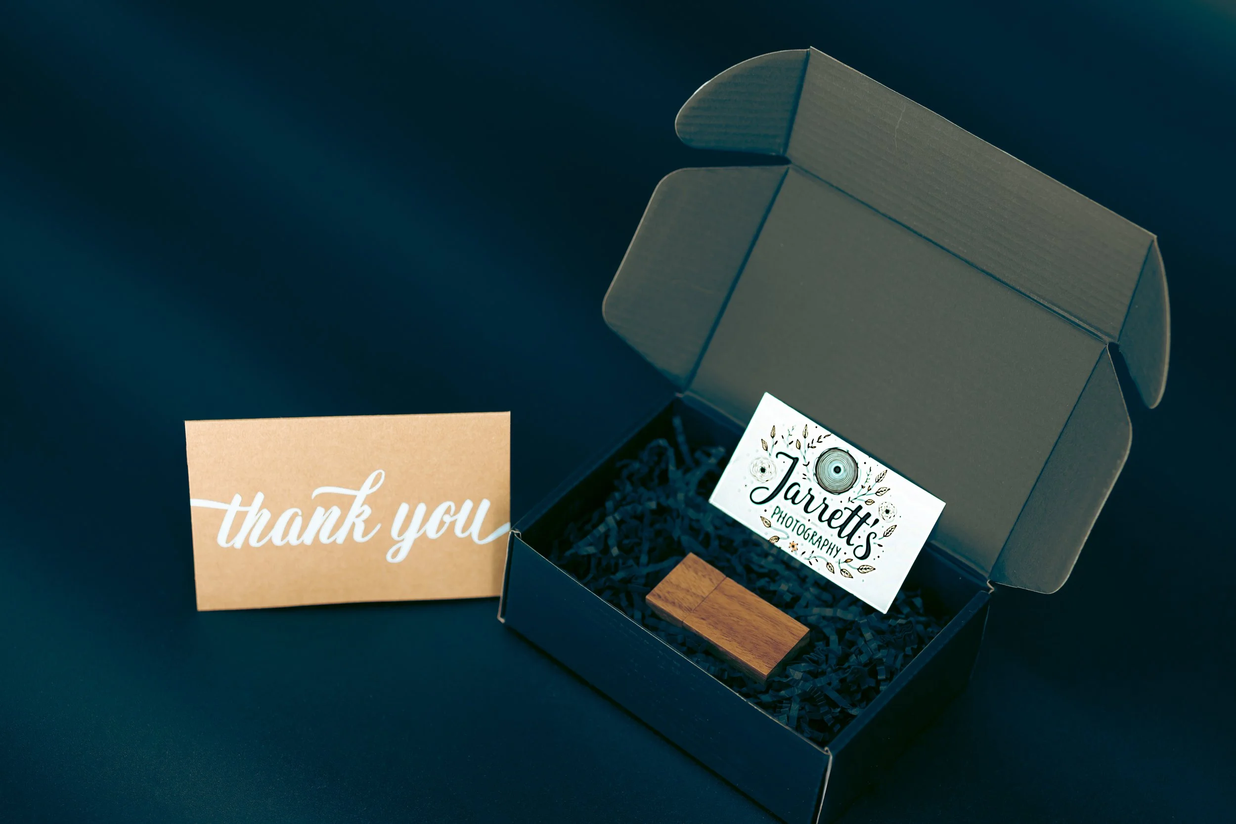 Open black gift box with shredded paper, a business card for Jarrett's Photography, a small wooden thumb drive, and a thank you card outside the box on a dark surface.
