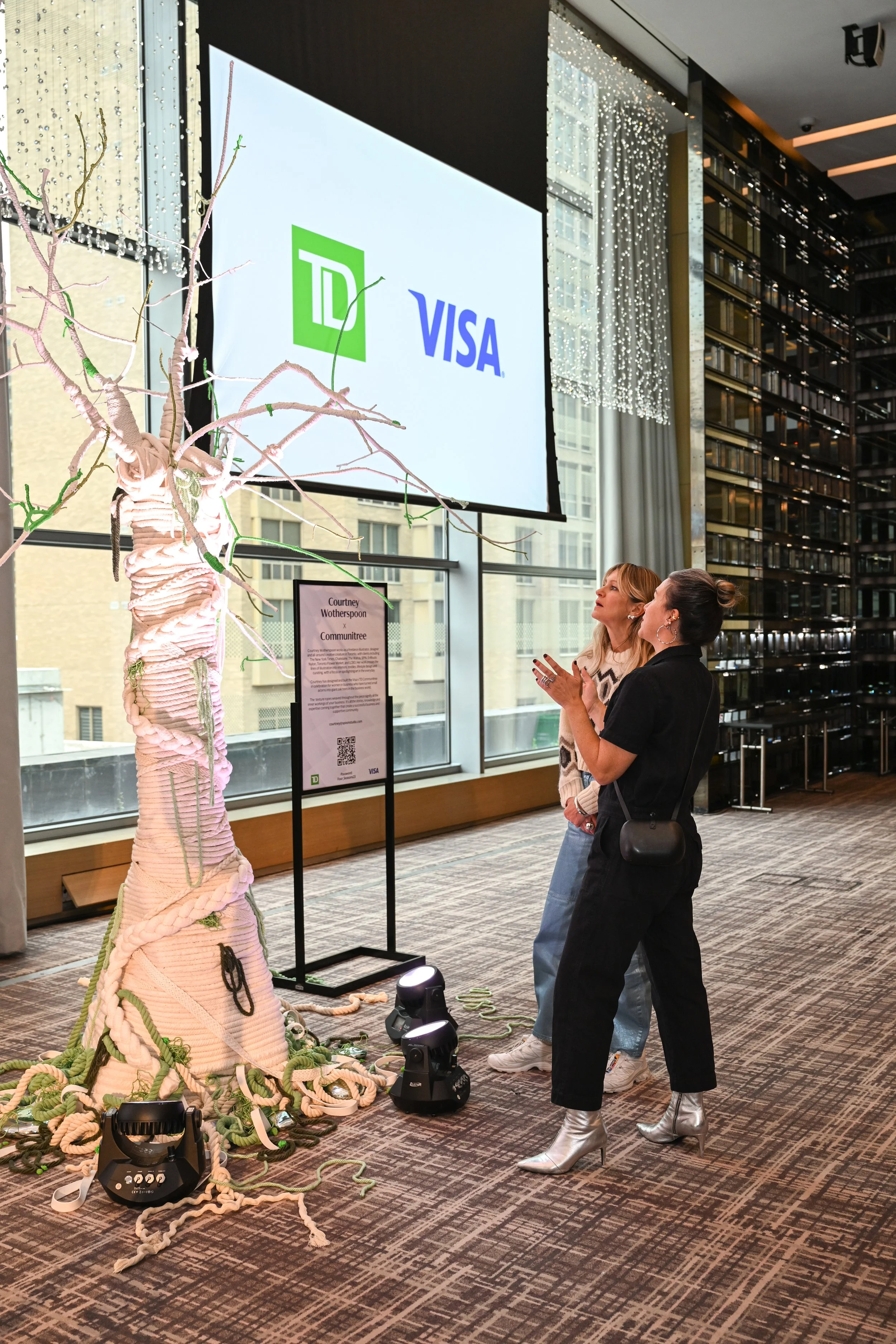 Two women in fashionable silver boots and casual clothing look at a tall art installation made of woven, tentacle-like ropes with small green accents, displayed inside a modern building with large windows and a digital screen showing logos of TD and Visa.