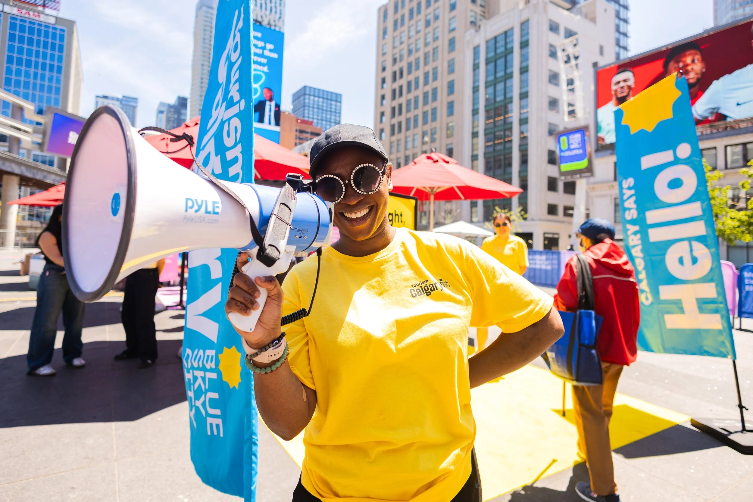 A woman in a yellow shirt with sunglasses holding a megaphone at a protest or rally in an urban area with tall buildings and banners.