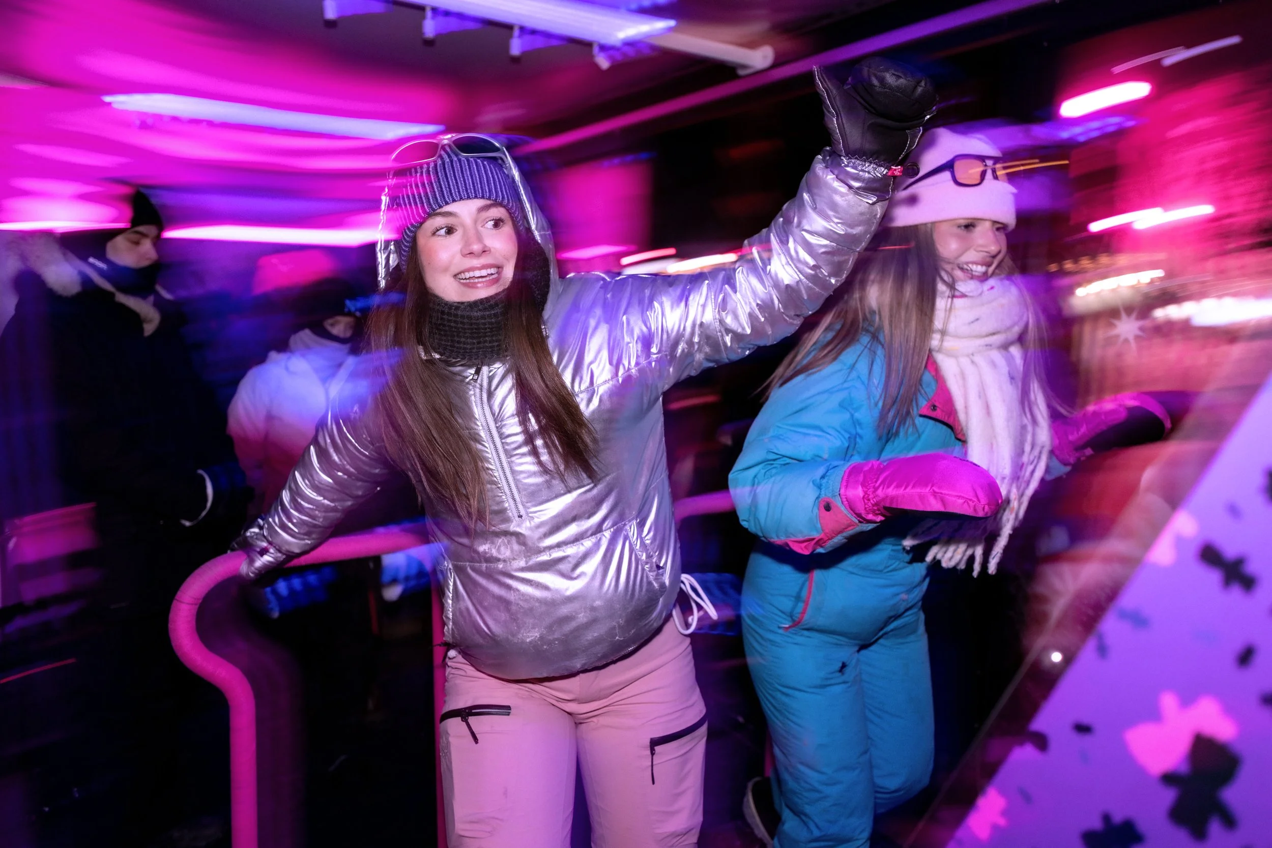 Two women in winter clothing, including jackets, gloves, hats, and scarves, are on a brightly lit ice skating rink, with one woman raising her arm and smiling, surrounded by colorful pink and purple lighting.