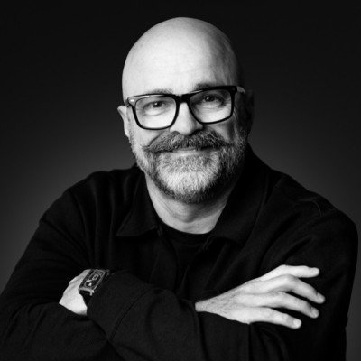 A black and white portrait of a bald man with glasses, smiling, wearing a black shirt with arms crossed.