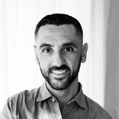 Black and white photo of a man with short dark hair and a beard, smiling at the camera, wearing a collared shirt.