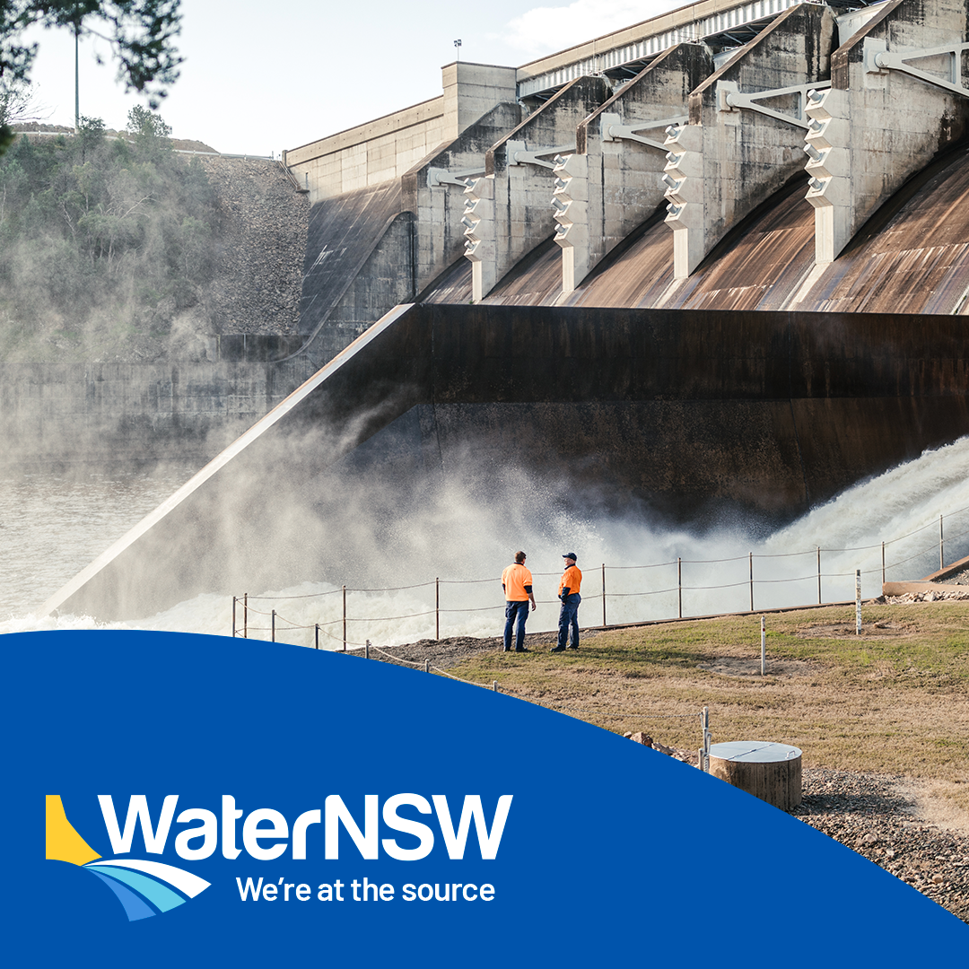 Two workers in orange shirts standing near a dam with water spilling over, with a logo for WaterNSW and the slogan 'We're at the source' in the foreground.