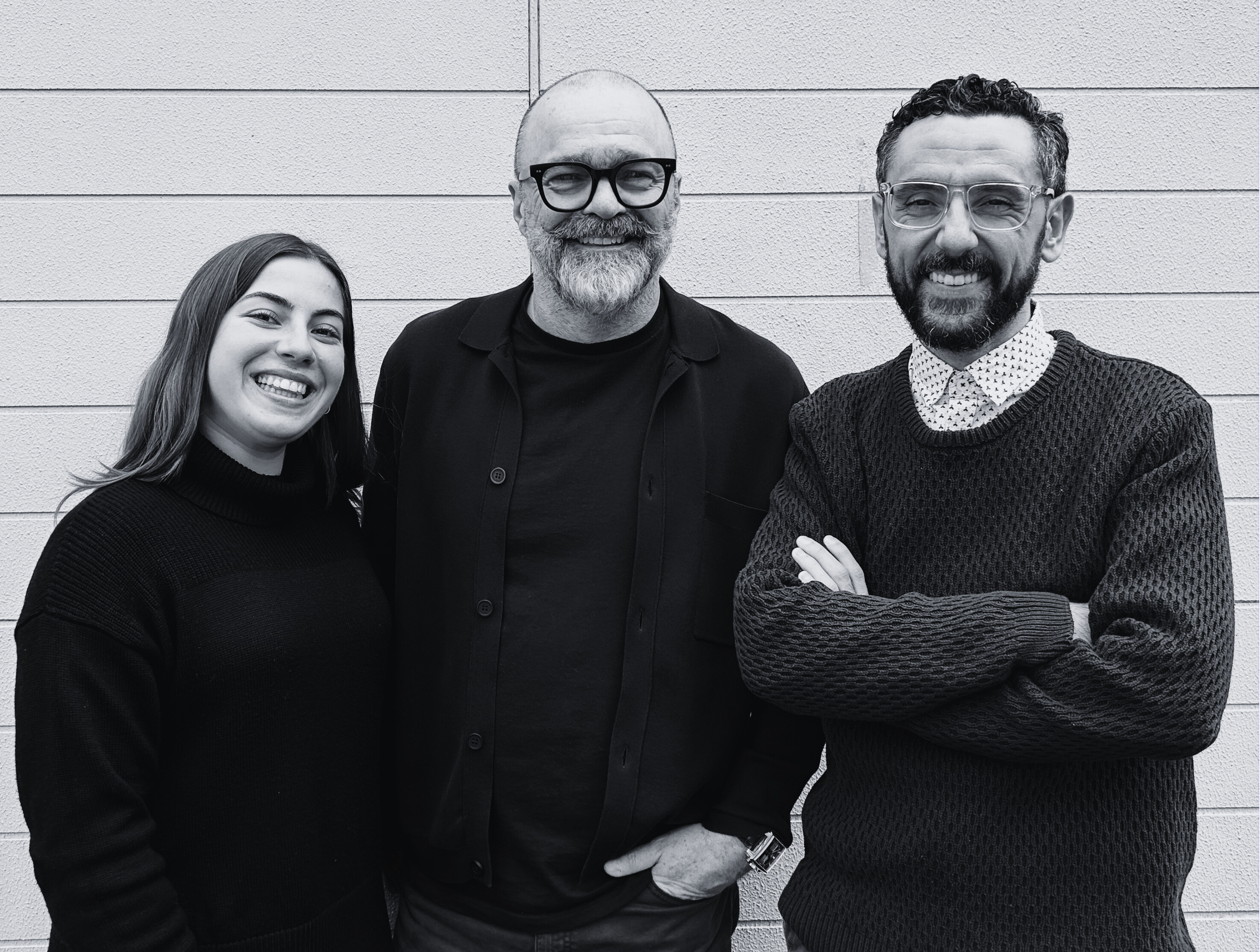 Three people smiling and posing against a white brick wall, with the person on the right crossing their arms.