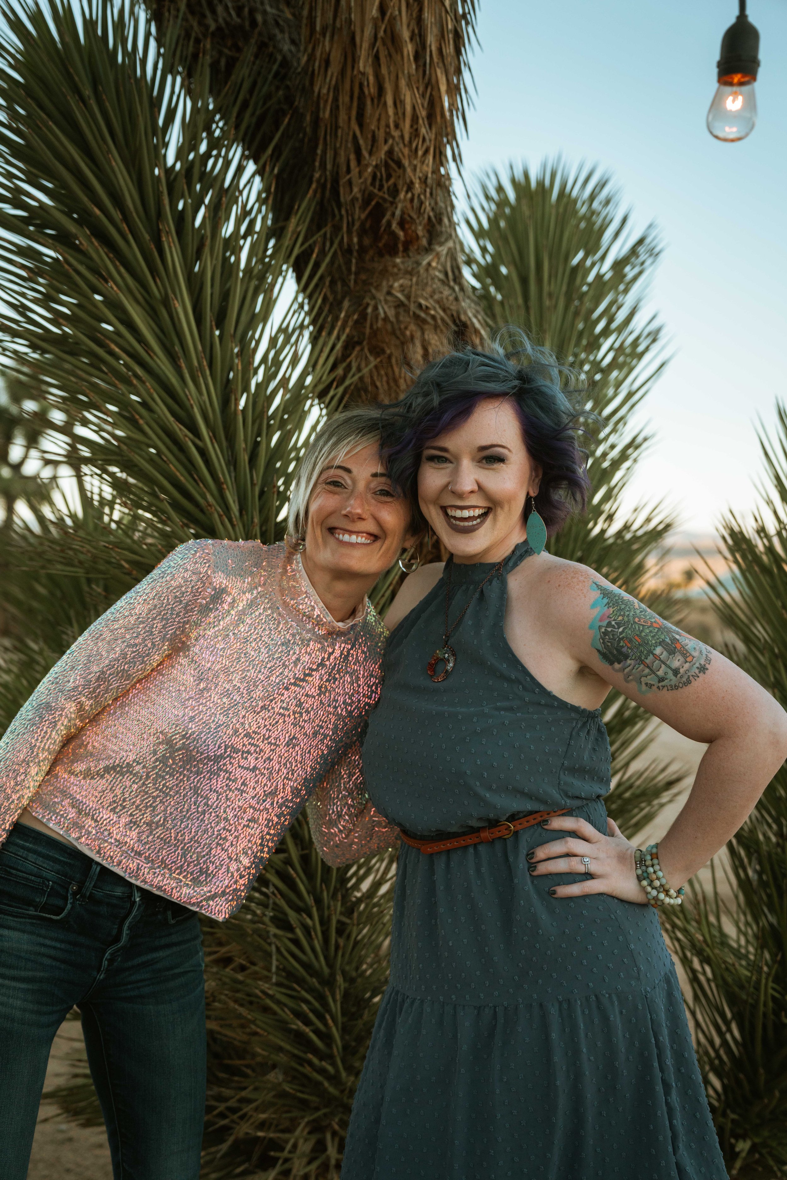 Two women smiling and posing for a photo outdoors in front of a large desert plant, with a sunset sky in the background.