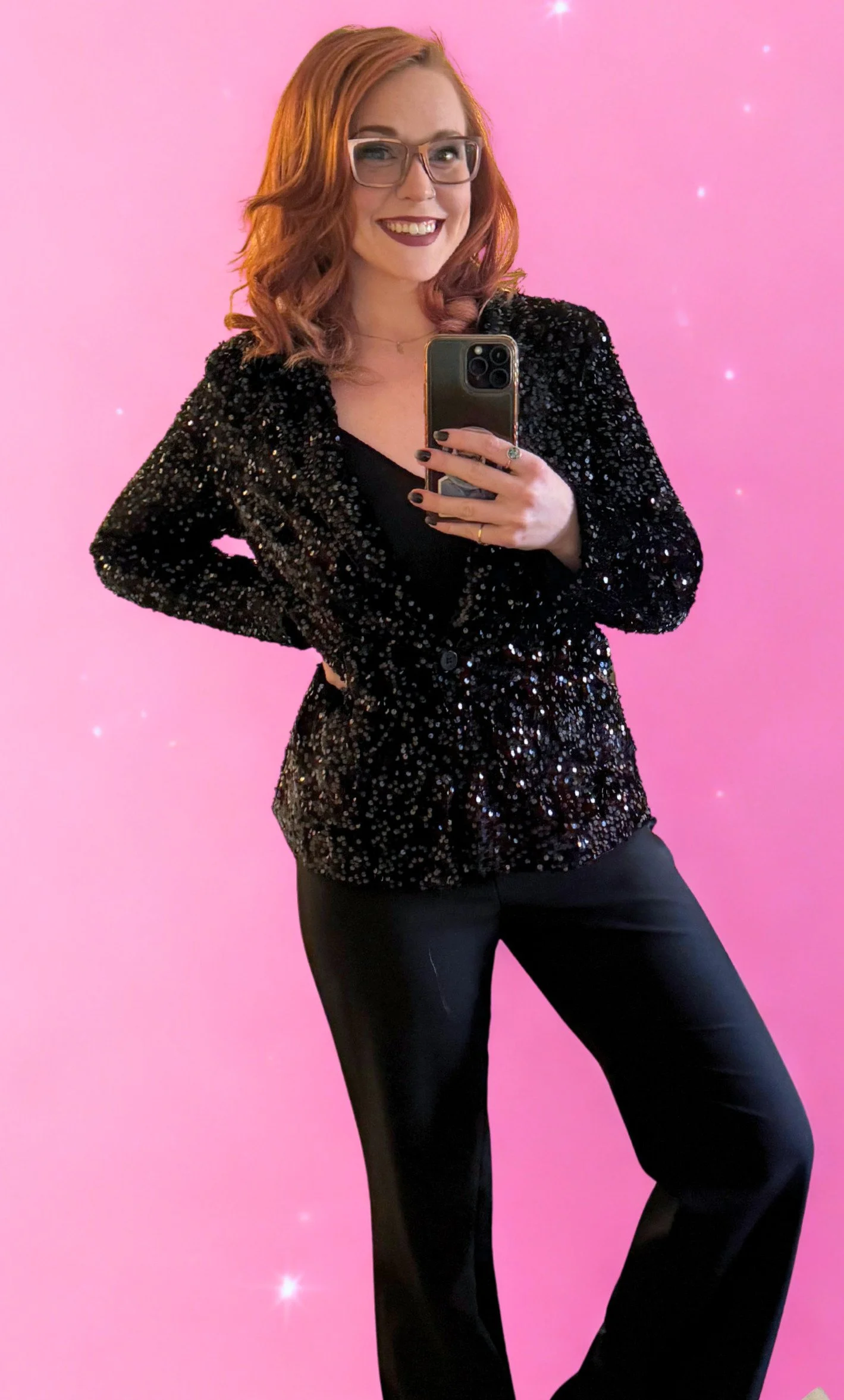 A woman with red hair, wearing glasses, a black sequined blazer, and black pants, taking a selfie in front of a pink background with small stars.