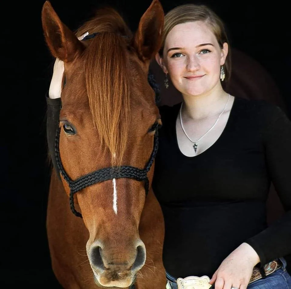 A young woman with blonde hair and a black top standing next to a brown horse with a white stripe on its face, both facing the camera.