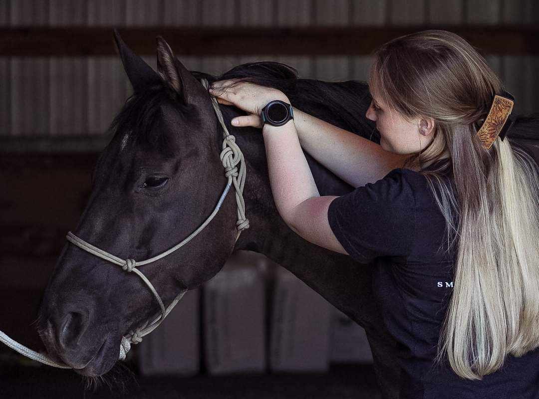 A woman with long blonde hair and wearing a black t-shirt gently holding a dark brown horse's head in a stable.