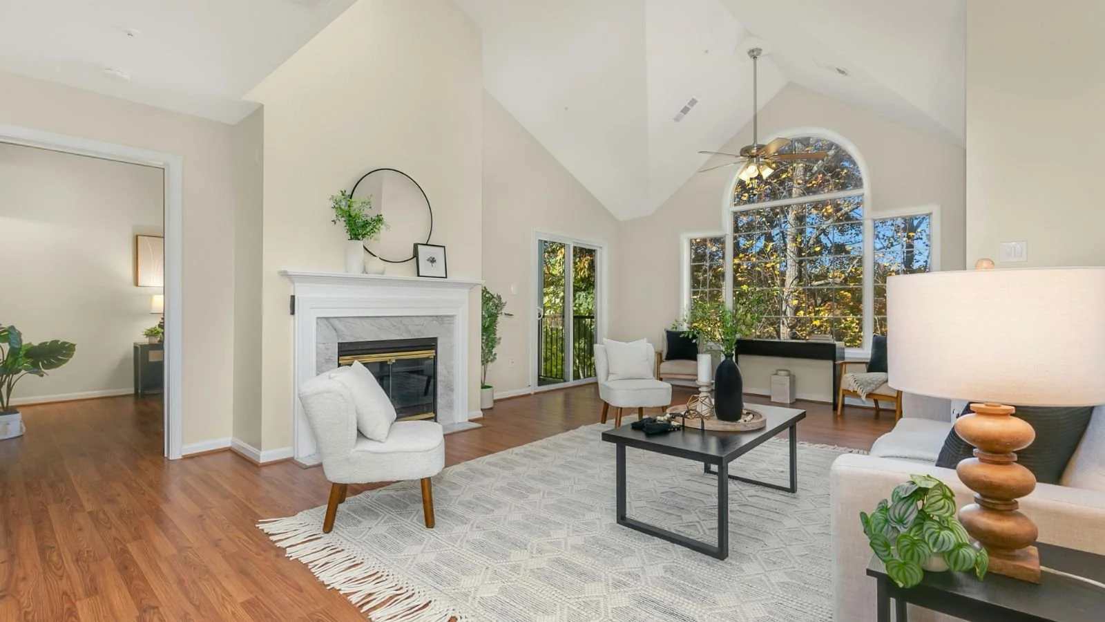 Bright living room with white walls, wooden floors, large arched window with trees outside, white fireplace, white and black furniture, coffee table, potted plants, ceiling fan, natural light.