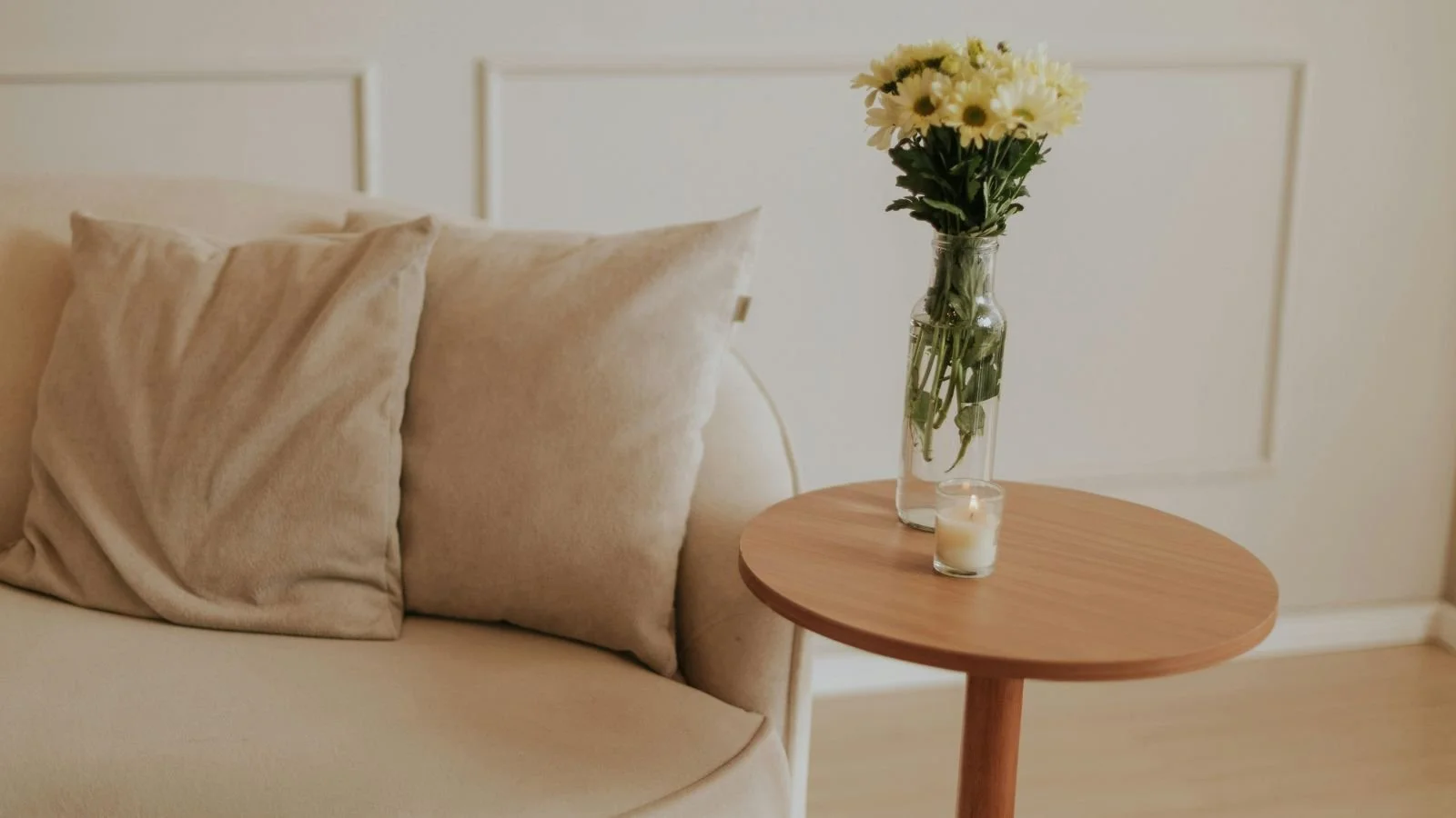 A cozy living room corner with a beige sofa, matching pillows, a wooden side table with a vase of white flowers, and a lit candle.