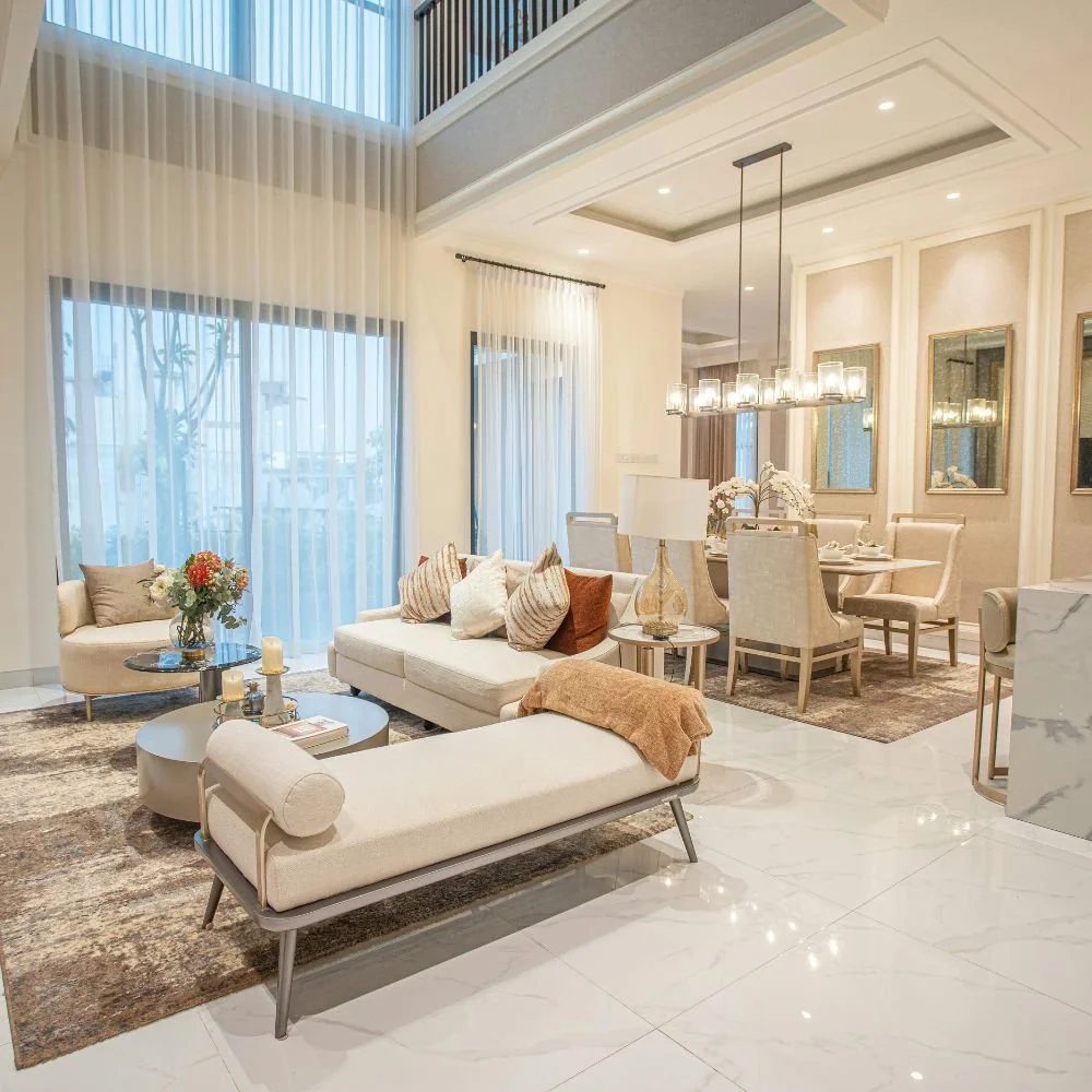 A bright, modern living and dining room with beige furniture, sheer curtains, a chandelier, and decorative mirrors.