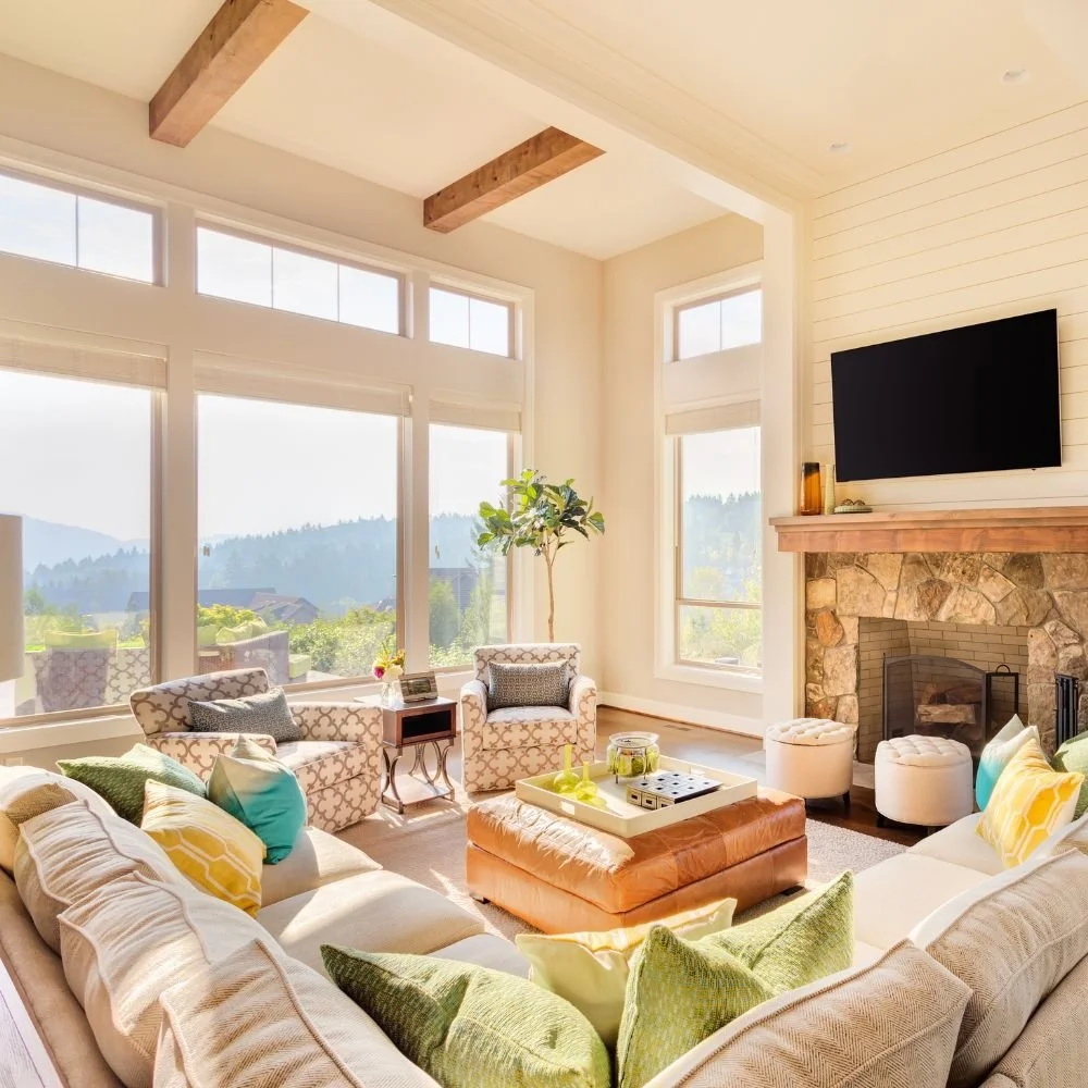 Bright living room with large windows overlooking mountains, a stone fireplace, a wall-mounted TV, and a variety of colorful and neutral sofas and chairs arranged around a central ottoman.