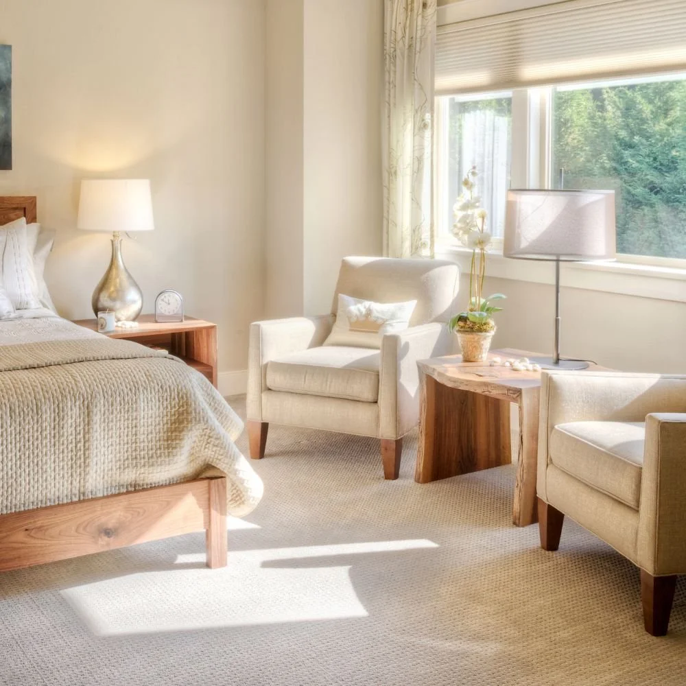 A cozy bedroom featuring a bed with white bedding and a wooden headboard, a nightstand with a lamp and small clock, a white armchair with a pillow, a tall glass lampshade, a side table with a potted orchid, and large windows with curtains allowing natural light to fill the room.