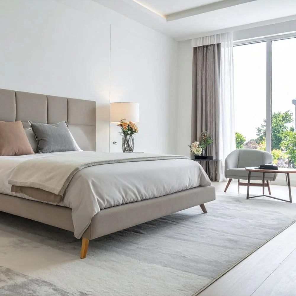 A modern bedroom with a large bed, neutral-colored bedding, and pillows. There is a nightstand with a lamp and flowers, a sitting chair and small table near a large window with curtains, letting in natural light and showing green trees outside.
