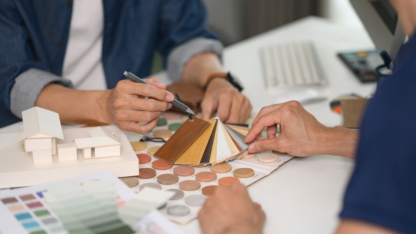 Two people discussing color and material options for interior design, with color swatches, wood samples, and architectural models on the table.