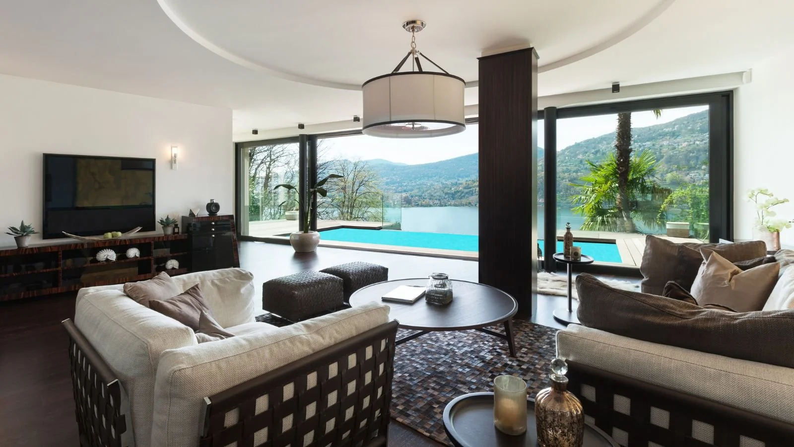 Living room with large windows overlooking a lake and mountains, featuring sofas, a coffee table, a television, and indoor plants.