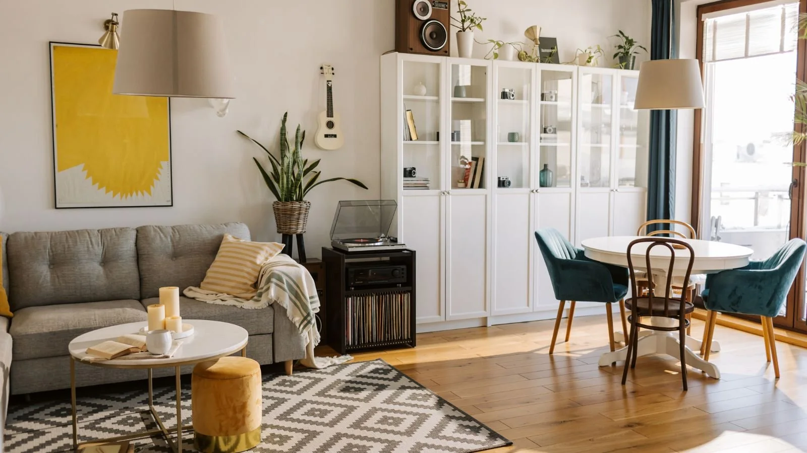 Living room with beige sofa, artwork, potted plant, record player, white cabinet, dining table with teal and brown chairs, wooden flooring, and large windows.