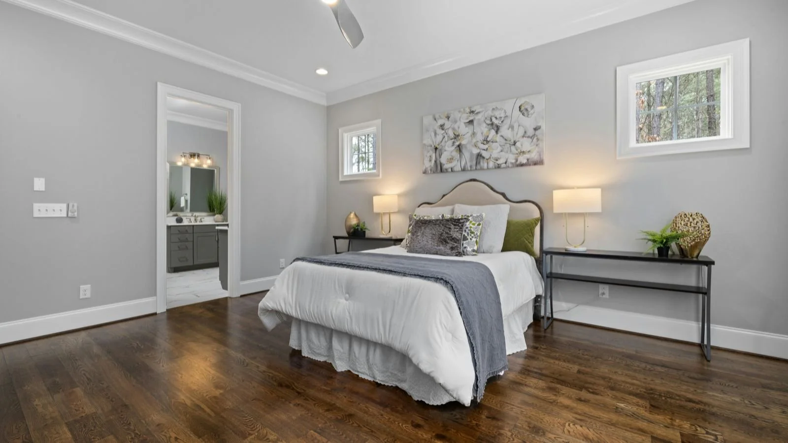A bedroom with gray walls, hardwood floors, a bed with white bedding and decorative pillows, two bedside tables with lamps, and a hallway leading to an en suite bathroom.