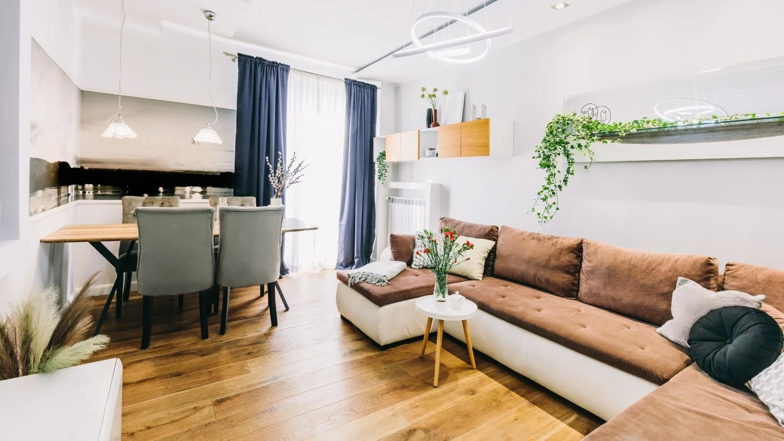 Living room with brown and beige sectional sofa, white side table with flowers, dining area with gray chairs and wooden table, large window with white and blue curtains, wall-mounted wooden shelves with plants, modern lighting fixtures