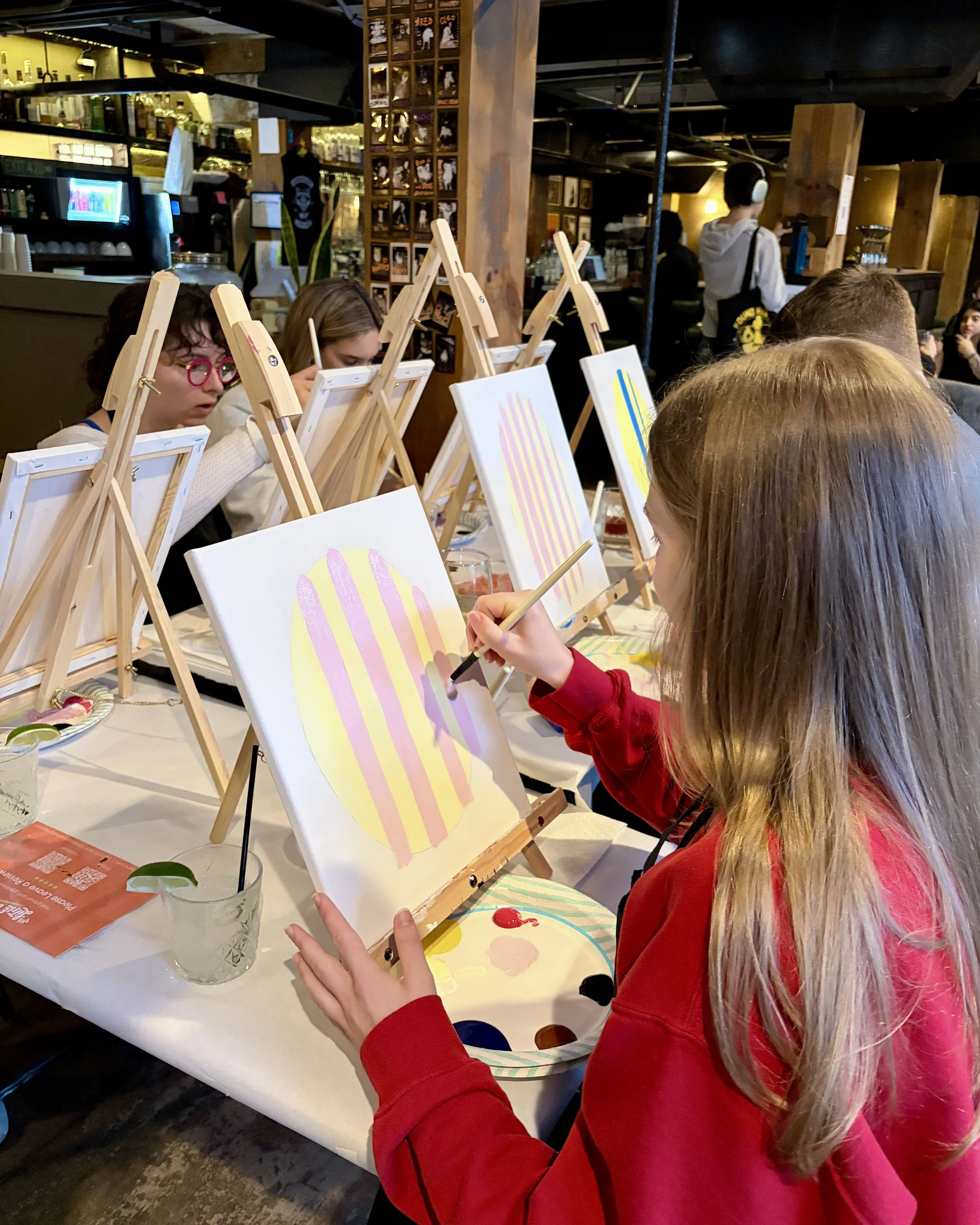 A girl with long hair in a red shirt paints stripes on her lemon painting in a coffee shop