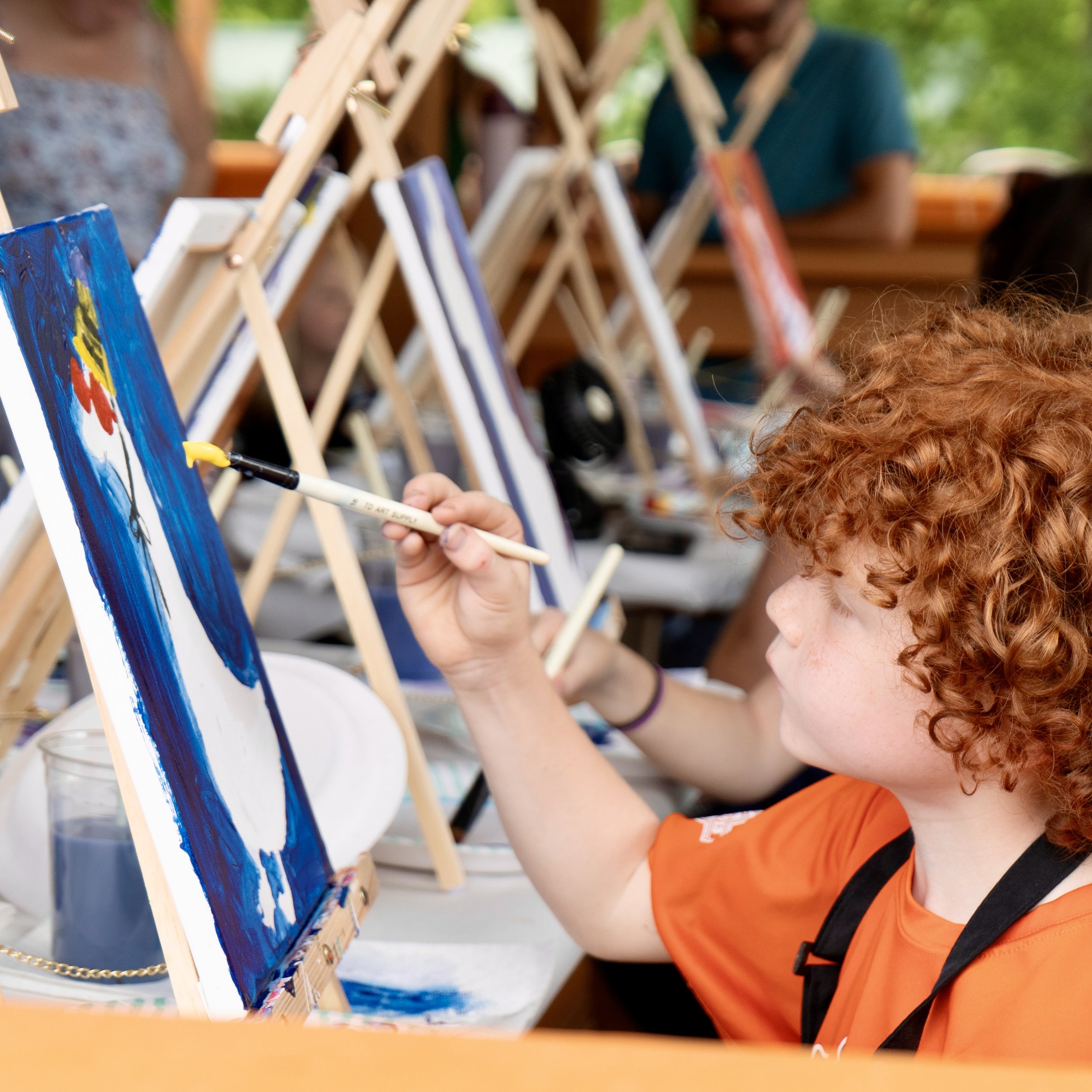 A boy with red curly hair holds arm out with yellow paint on brush to paint a canvas