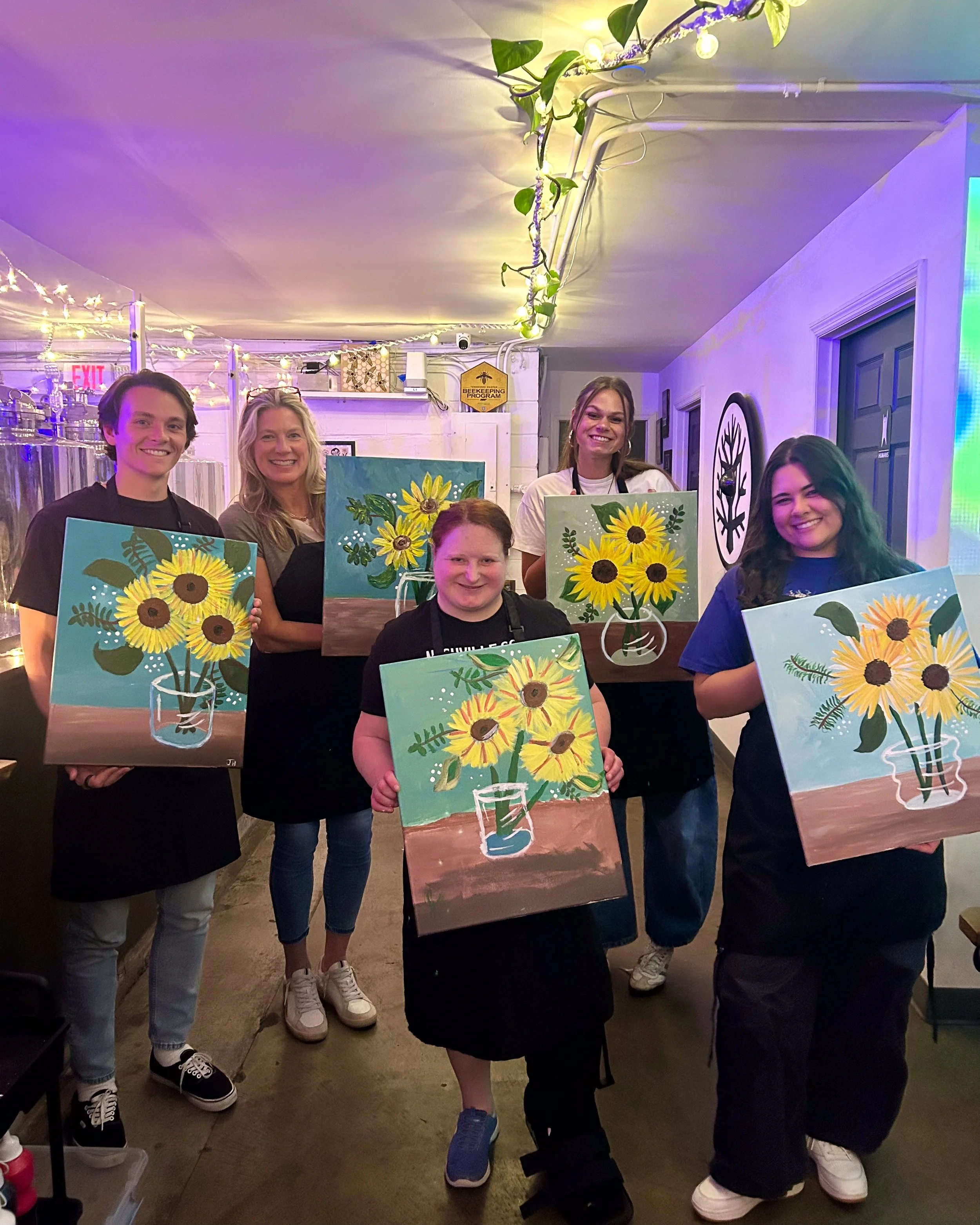 Group of people holding up their finished paintings of sunflowers in a vase in a colorfully lit indoor space