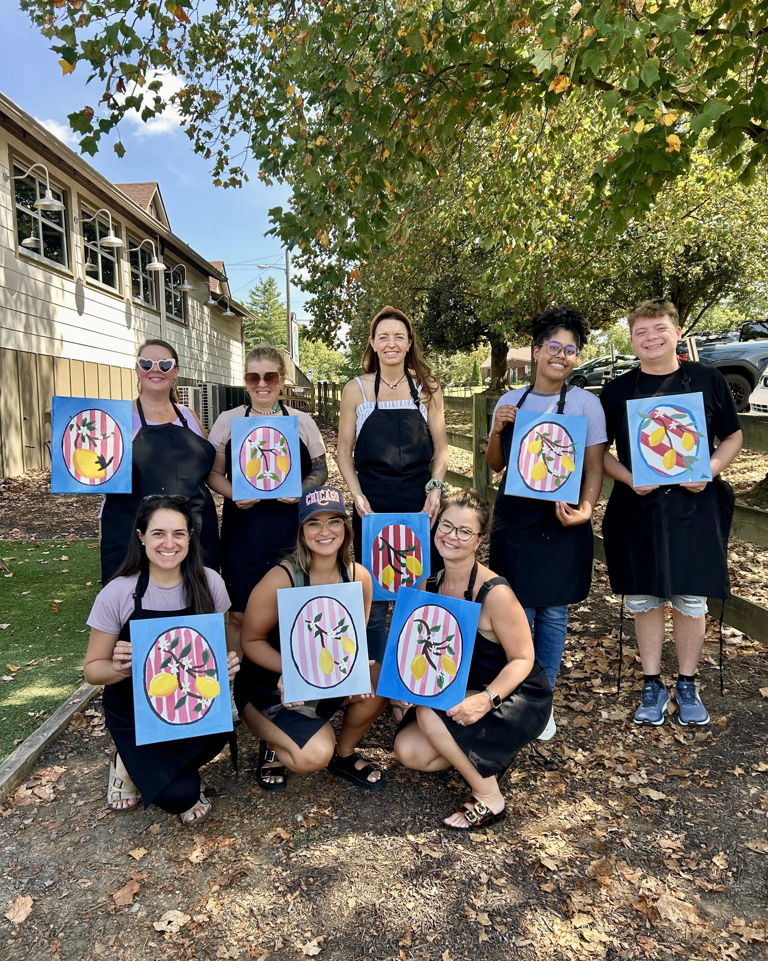 Group of eight adults in aprons holding lemon paintings and smiling at the camera outside on a sunny day under a green tree