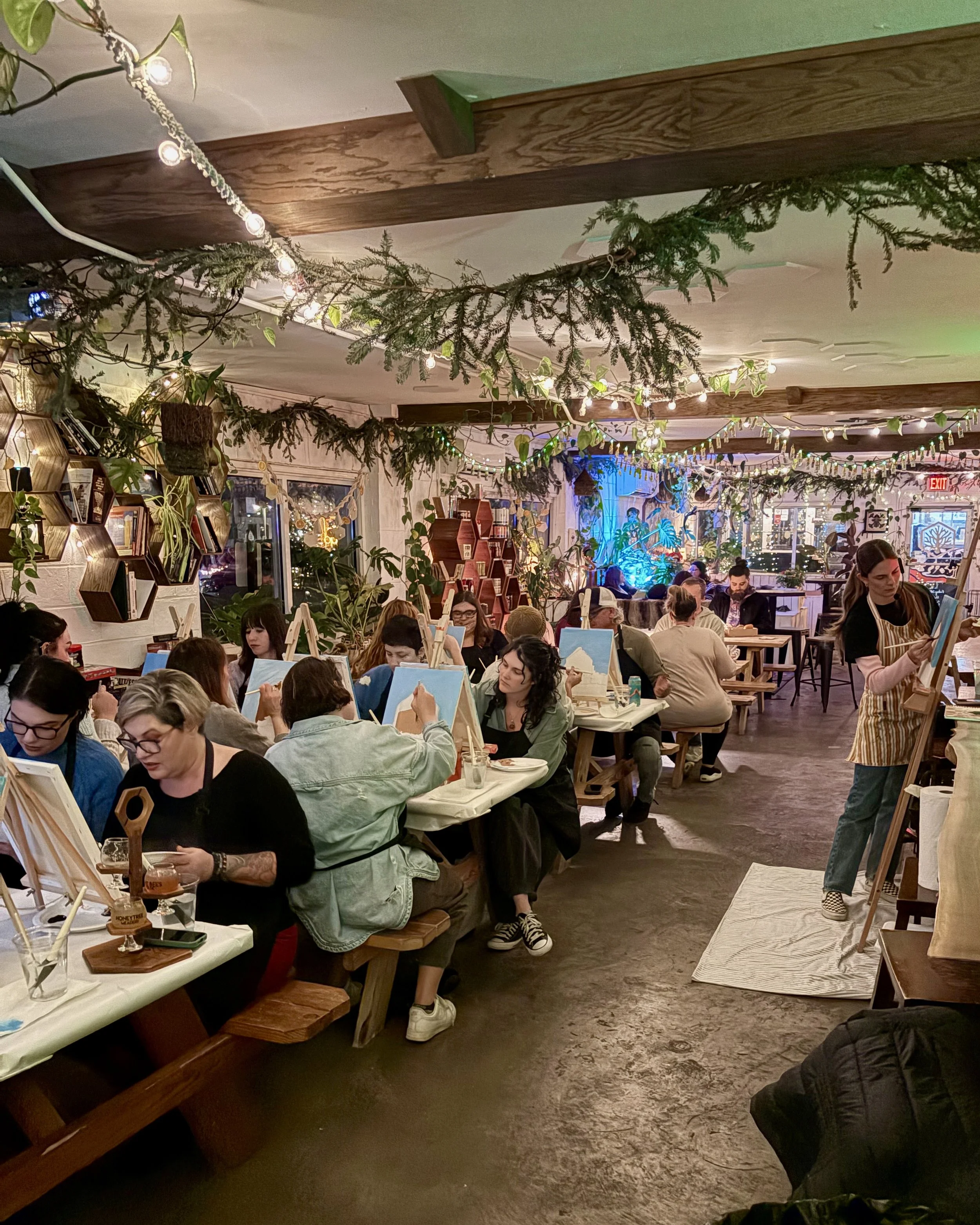A large group of people paint gingerbread houses indoors surrounded by twinkling lights and garland during the holidays
