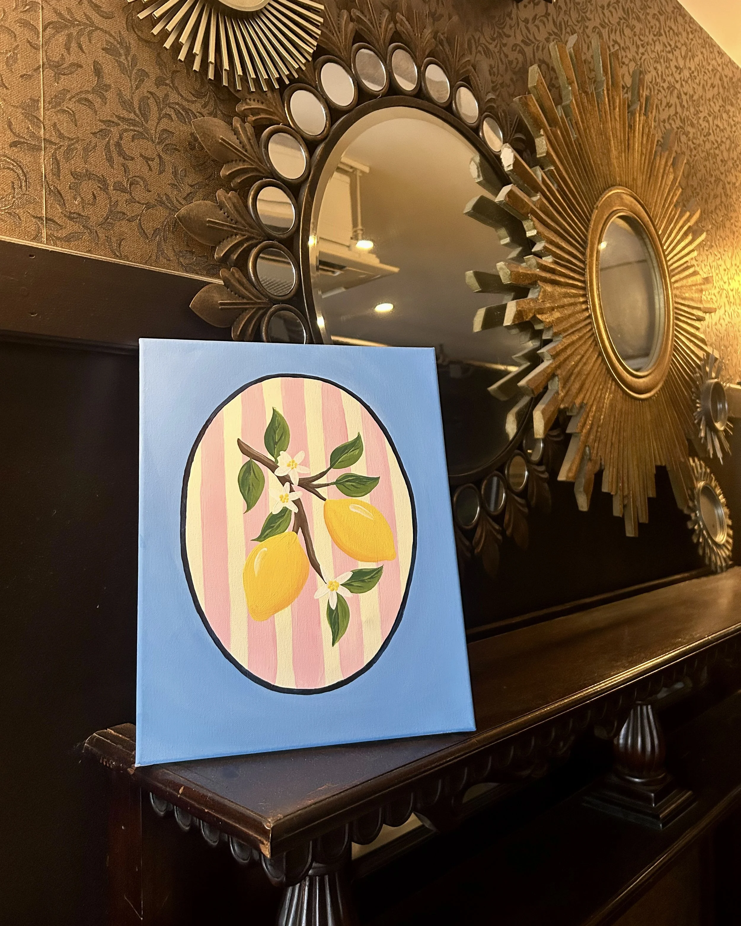 Up close photo of two lemons on a branch with flowers with a pink stripe background inside an oval on a light blue background