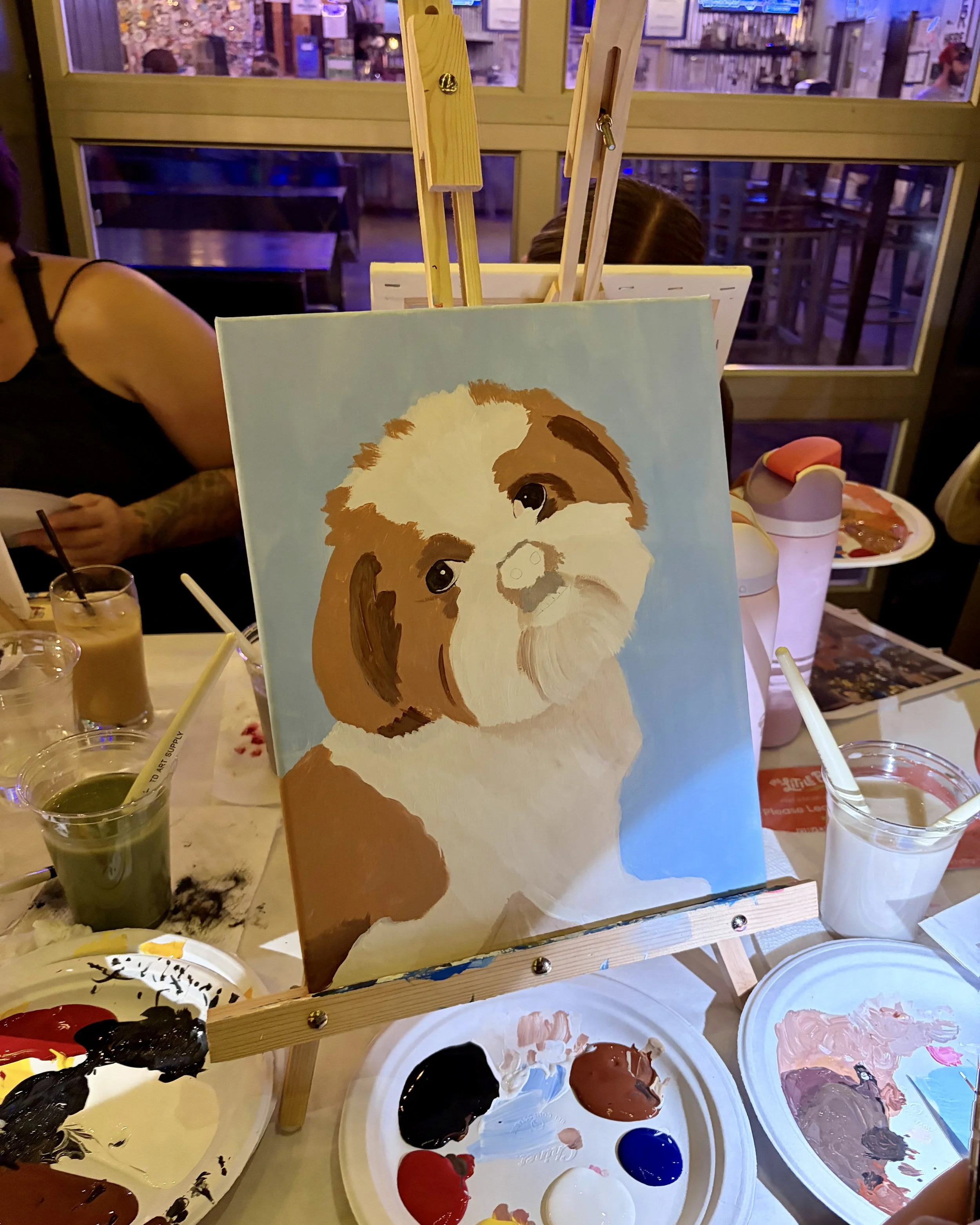 A brown and white Shih Tzu painting in progress with paint and art supplies