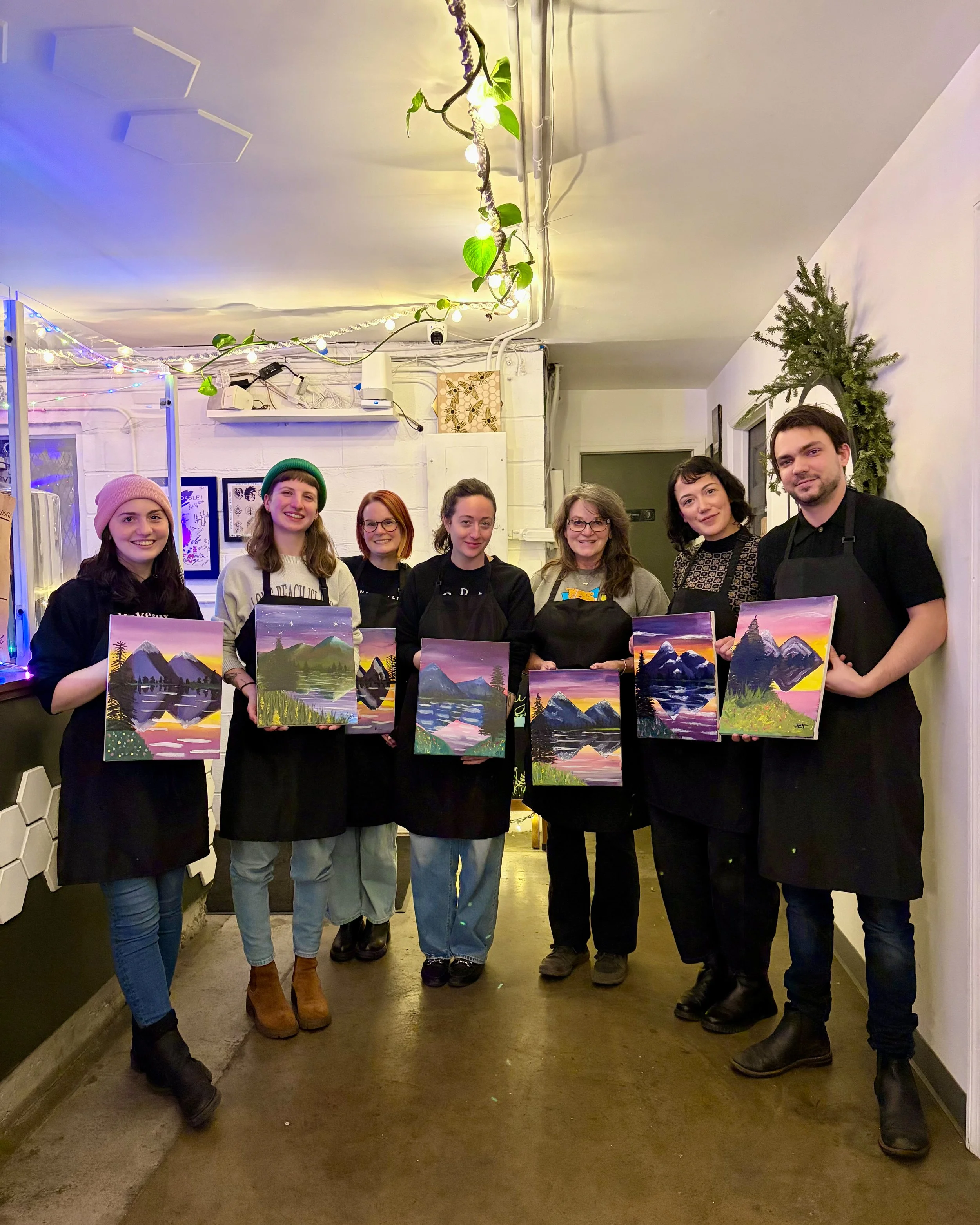 A group of people pose inside with string lights above as they hold up finished paintings of a mountain scene