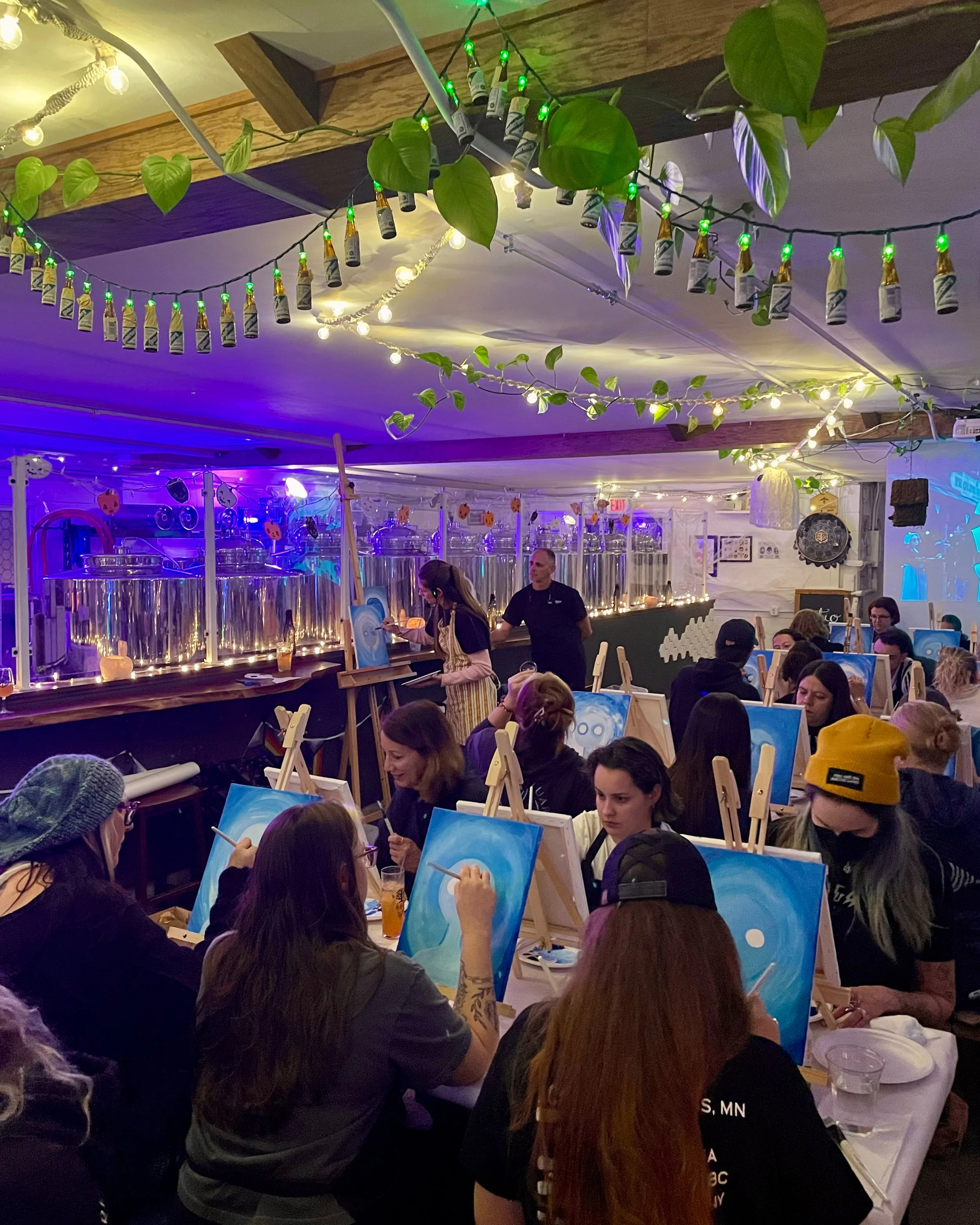 A large group of people paints spooky self portraits indoors with string lights overhead