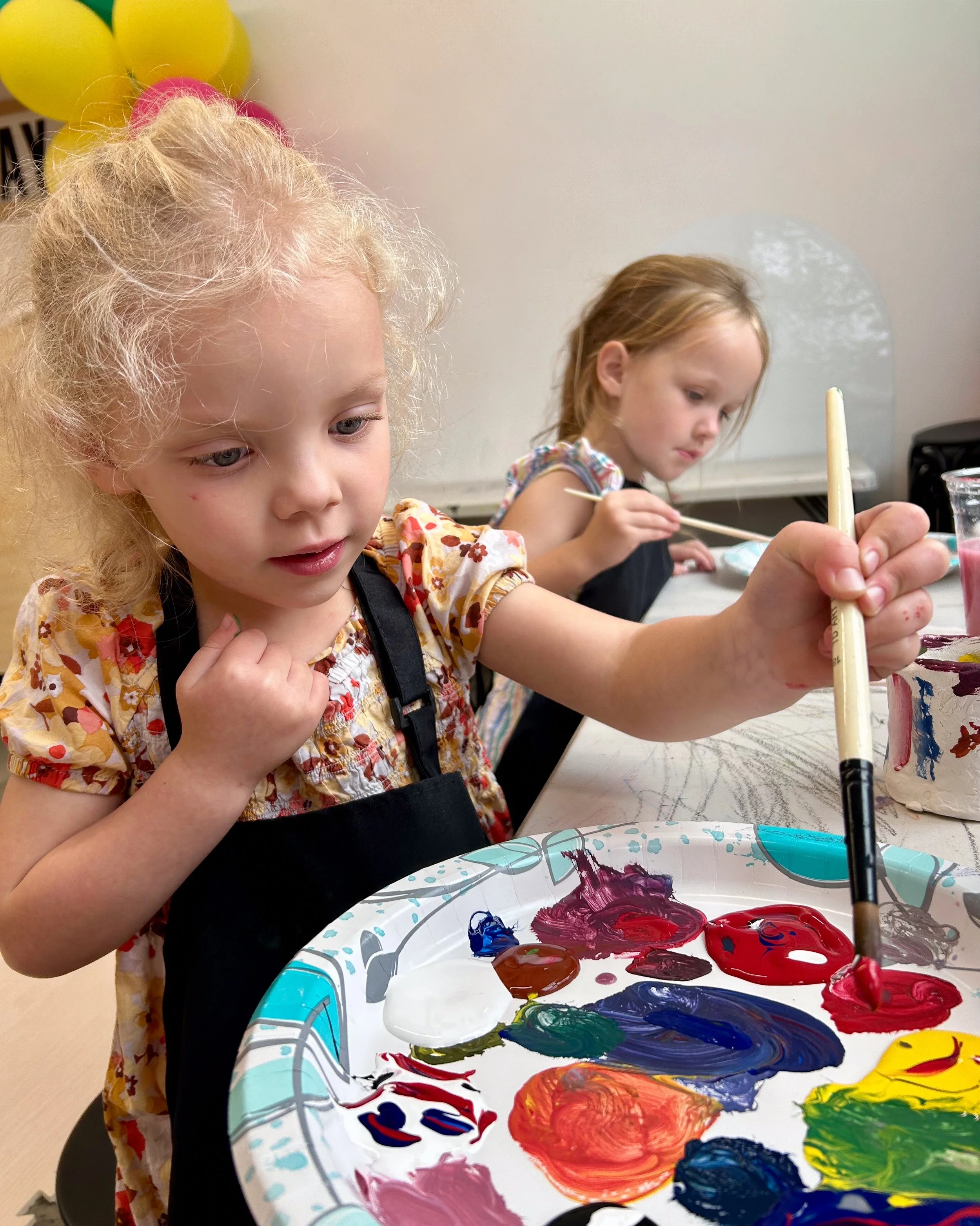 A little girl holding a paintbrush mixes colors on her paint palette