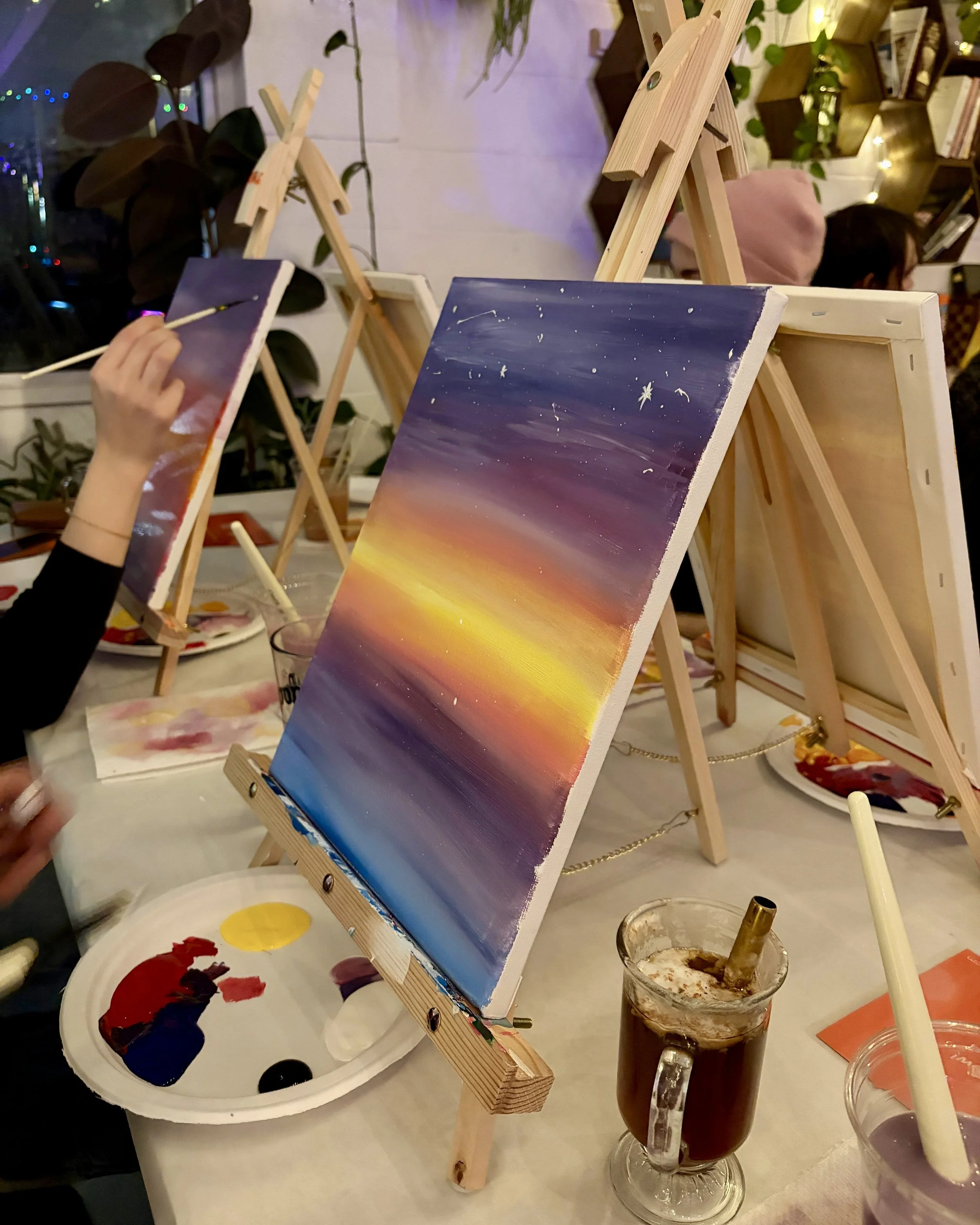 Up close photo of a sunset mountain scene in progress surrounded by painting supplies