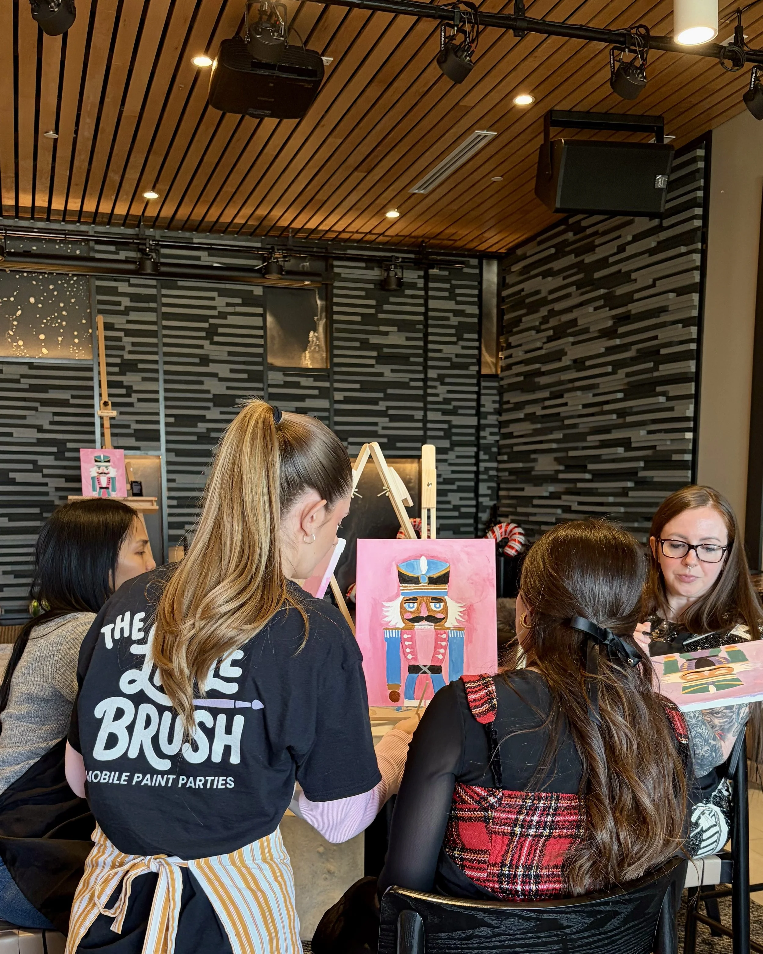 The back of an instructor in a Little Brush shirt helping a student with their pink nutcracker painting