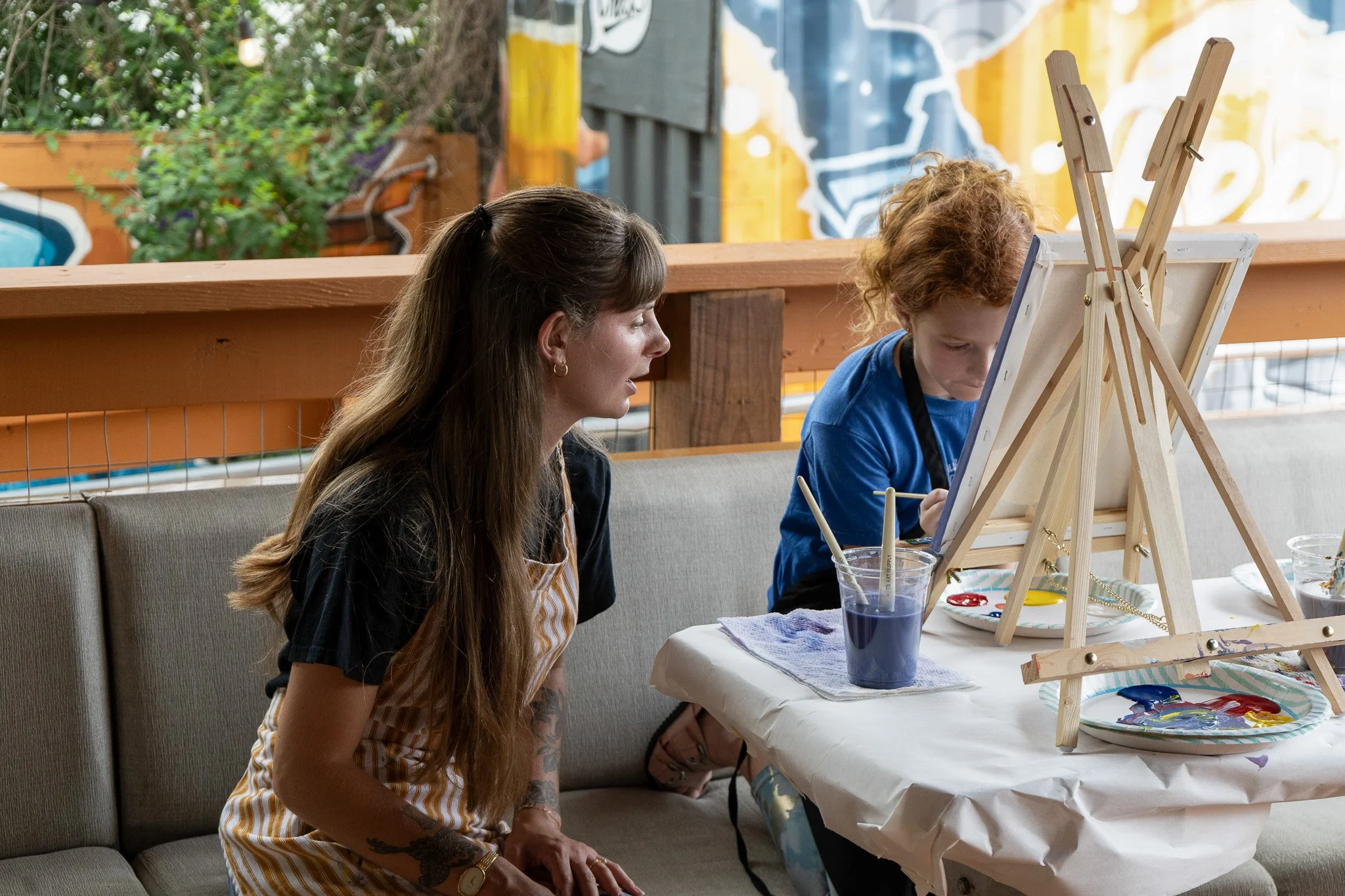 An instructor with long hair sits with a young girl and helps her paint her canvas on a patio