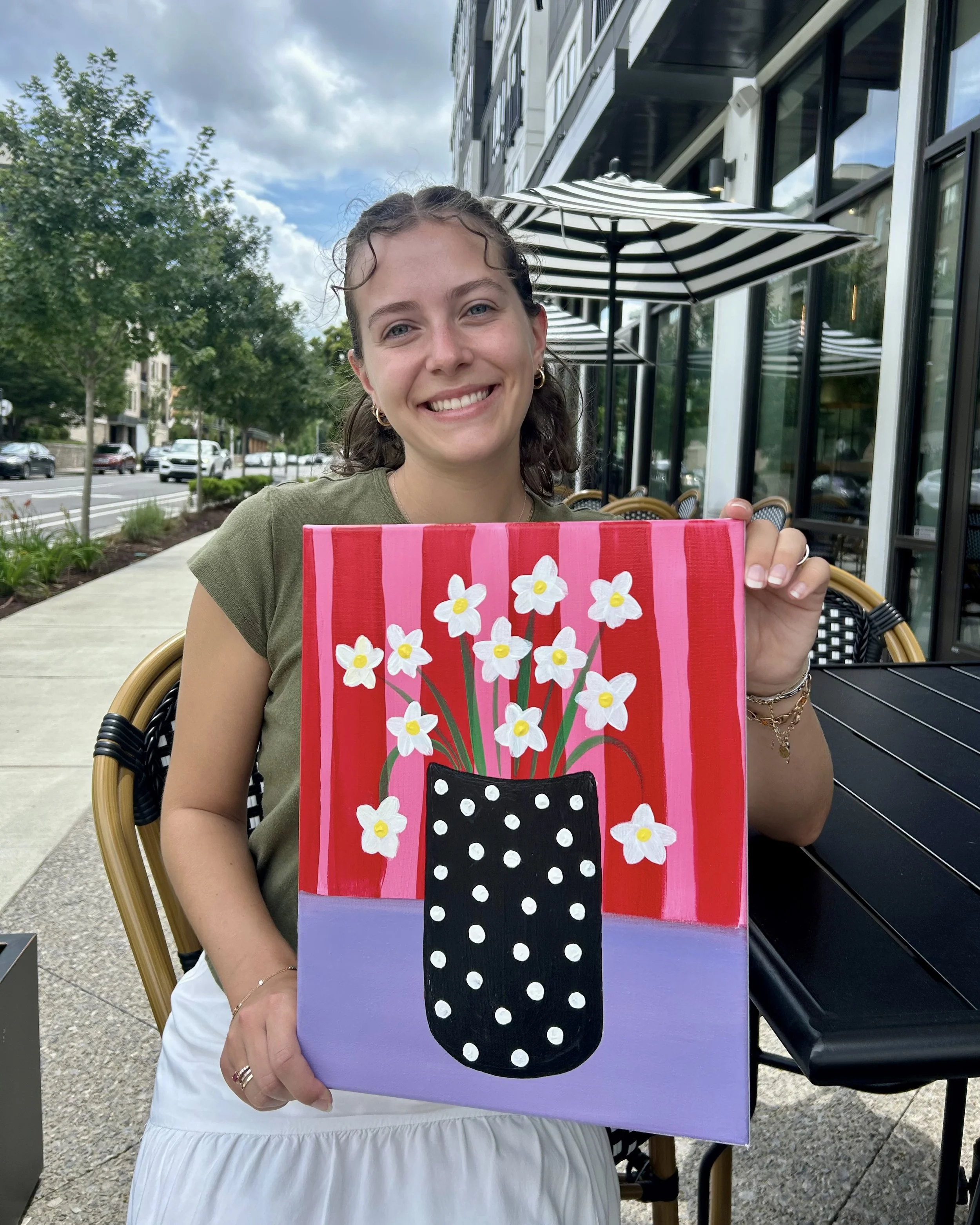A woman with brown curly hair smiles and holds up a pink and purple stripe painting with white flowers in a black polka dot vase