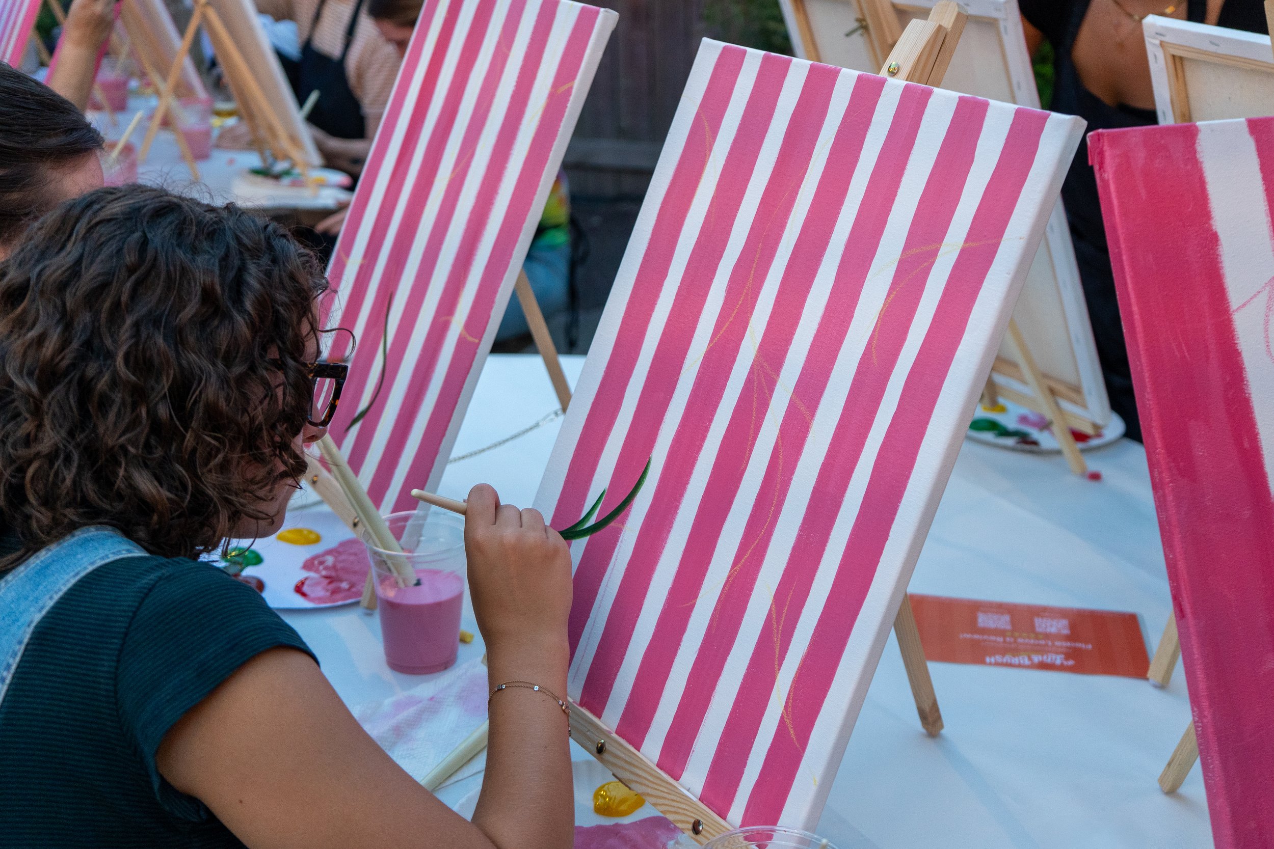 A girl with curly brown hair and glasses concentrates on painting tomato vines on her canvas