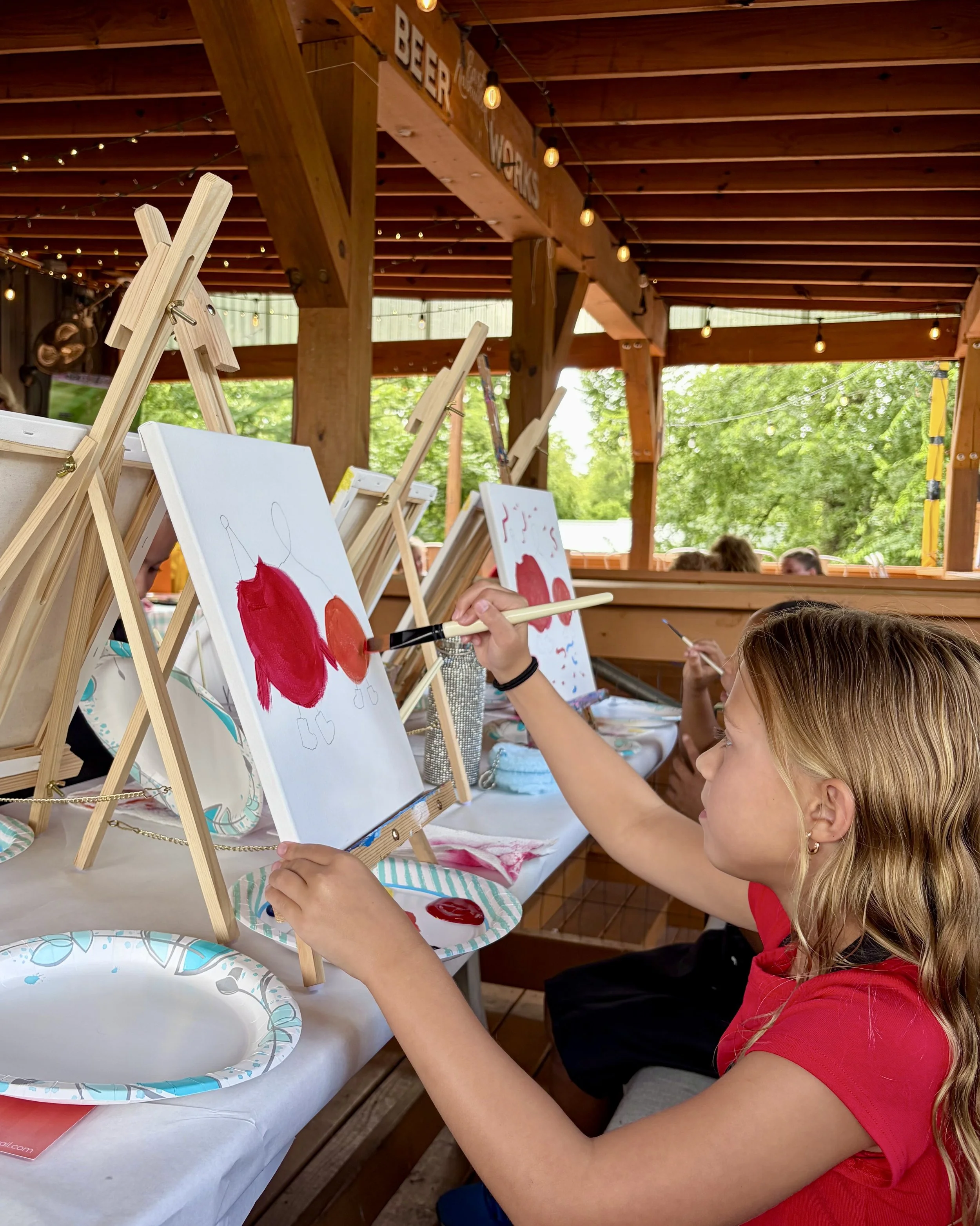 Little girl with wavy hair and a red shirt painting two tomatoes on a canvas outside