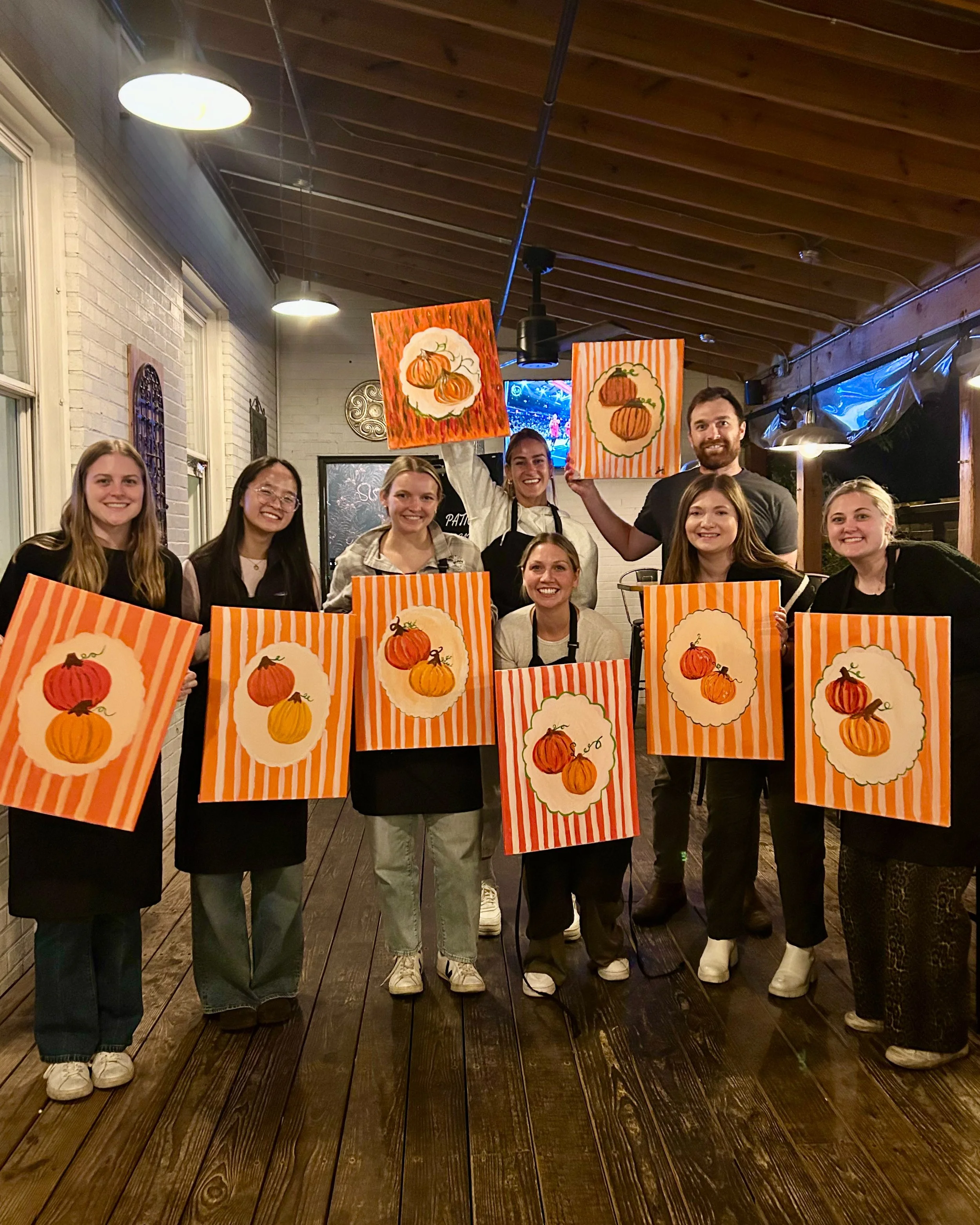 A group of people on a patio pose and hold up their finished pumpkin paintings