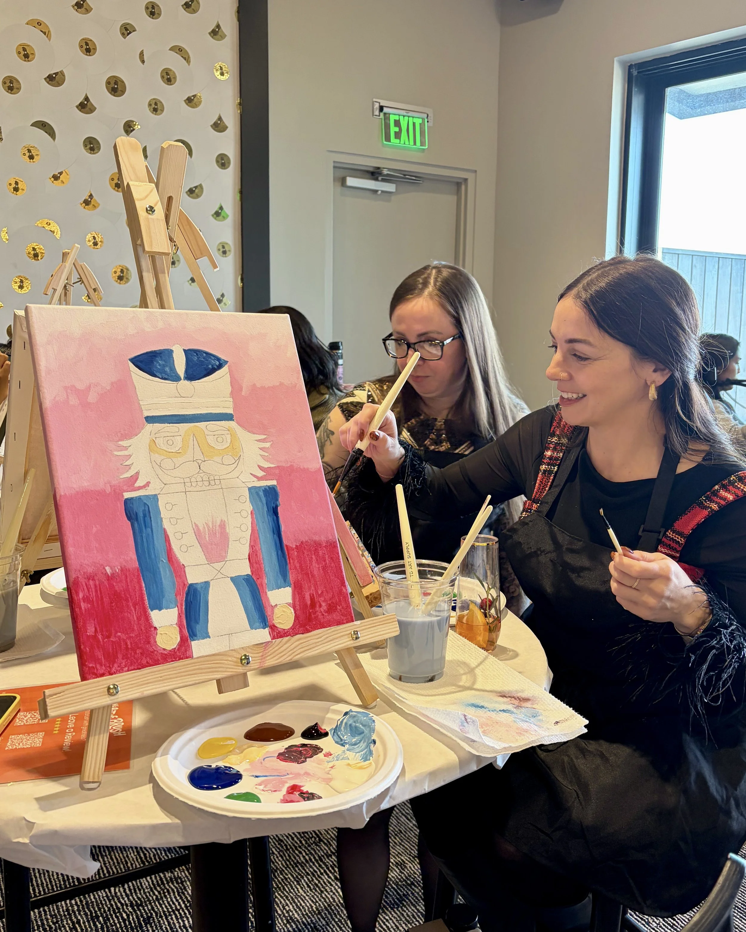 A woman with dark hair smiles and paints in the background while a pink and blue nutcracker painting is in the foreground