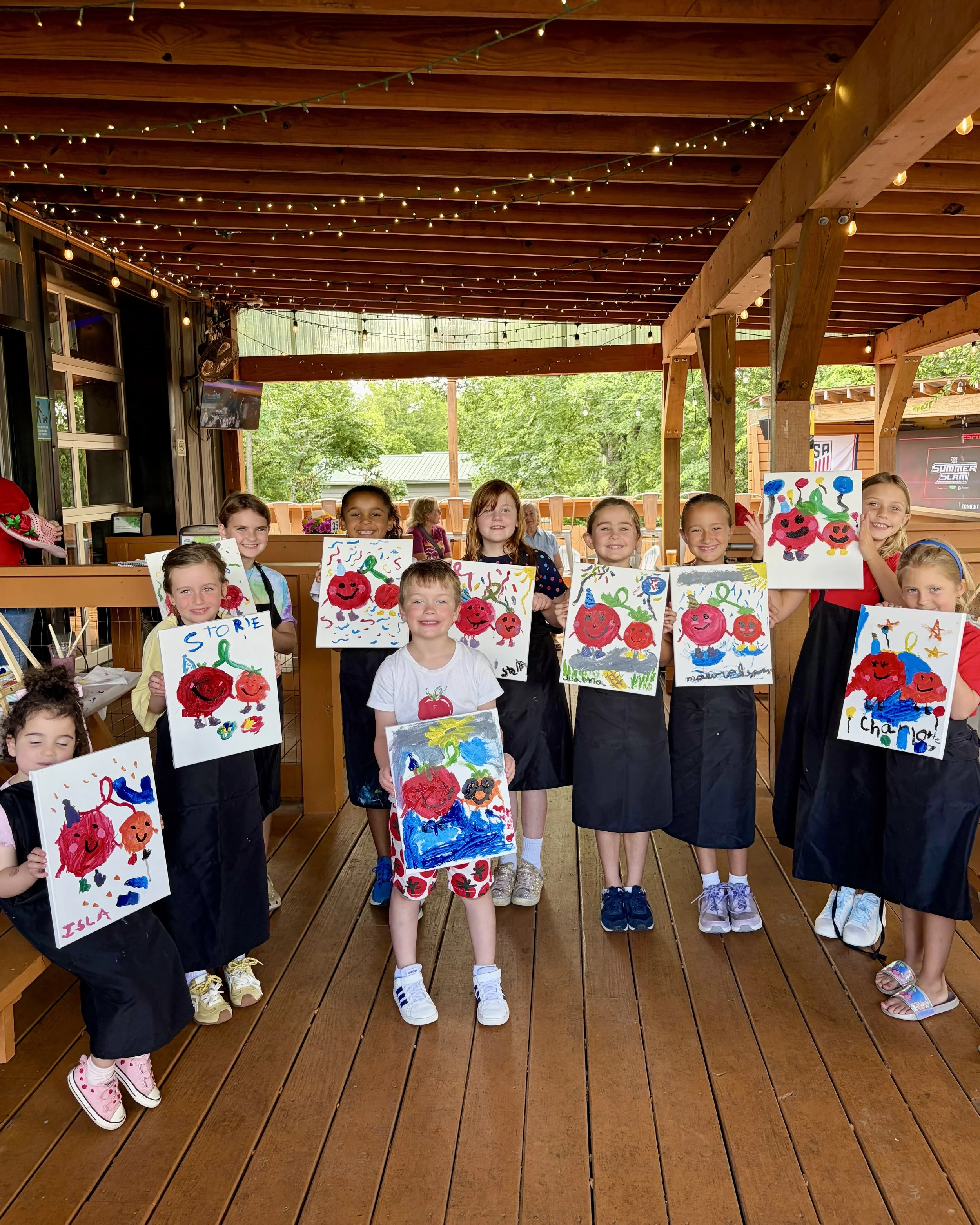 Group of children standing on a patio holding their tomato paintings on display and smiling at the camera