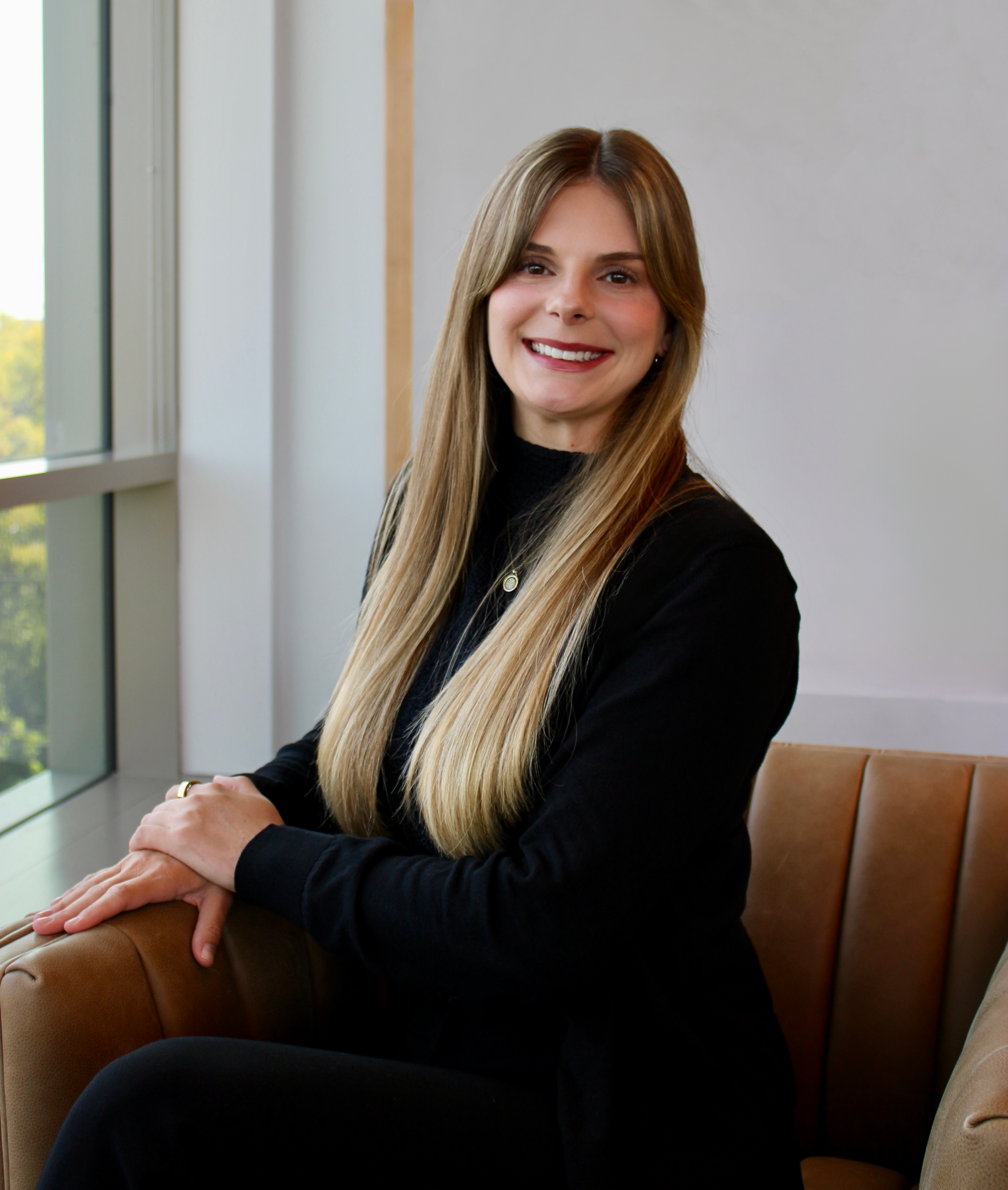 A girl with long blonde hair sits in a chair and smiles at the camera in a black outfit