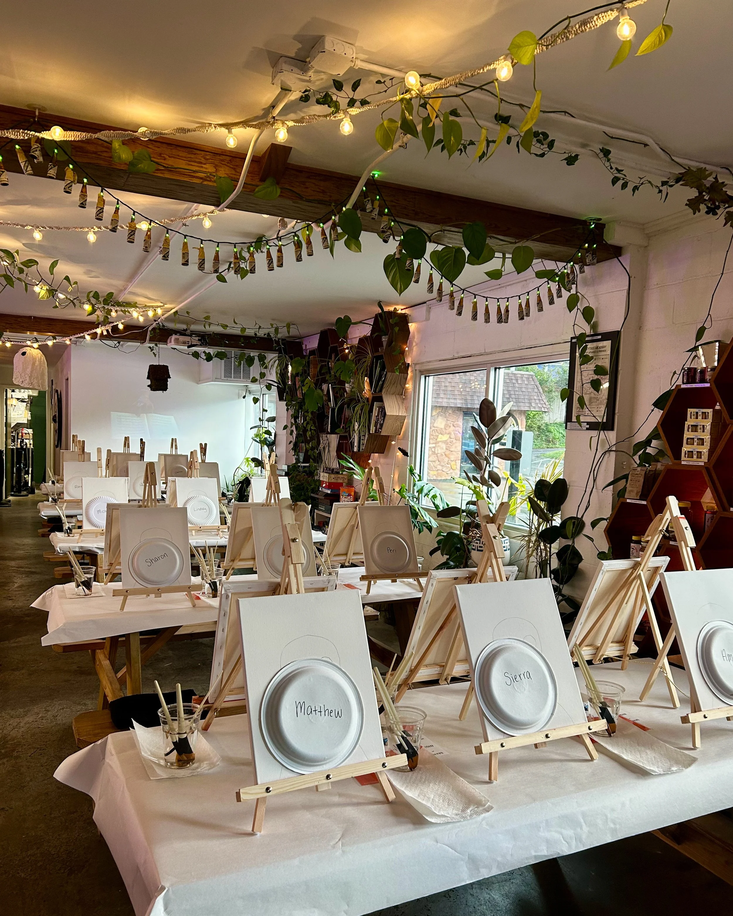 Multiple tables indoors with string lights above and canvases, easels, paper plates with names, and paintbrushes